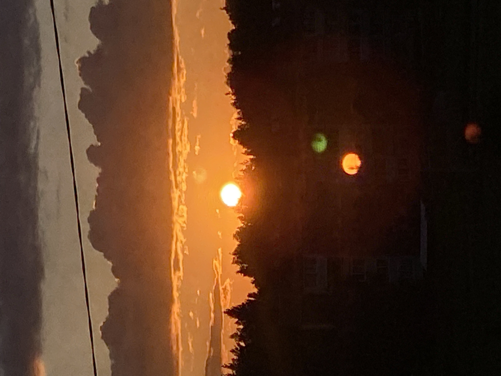 A sunset is depicted in the sky over a tree line, with a portion of a building visible in the foreground. The sky is filled with orange and gray clouds. A thin black wire stretches across the top of the image. Several blurred orbs of light are present in the lower portion of the image, appearing to be lens flares. The scene is likely from Марковский, Russia.