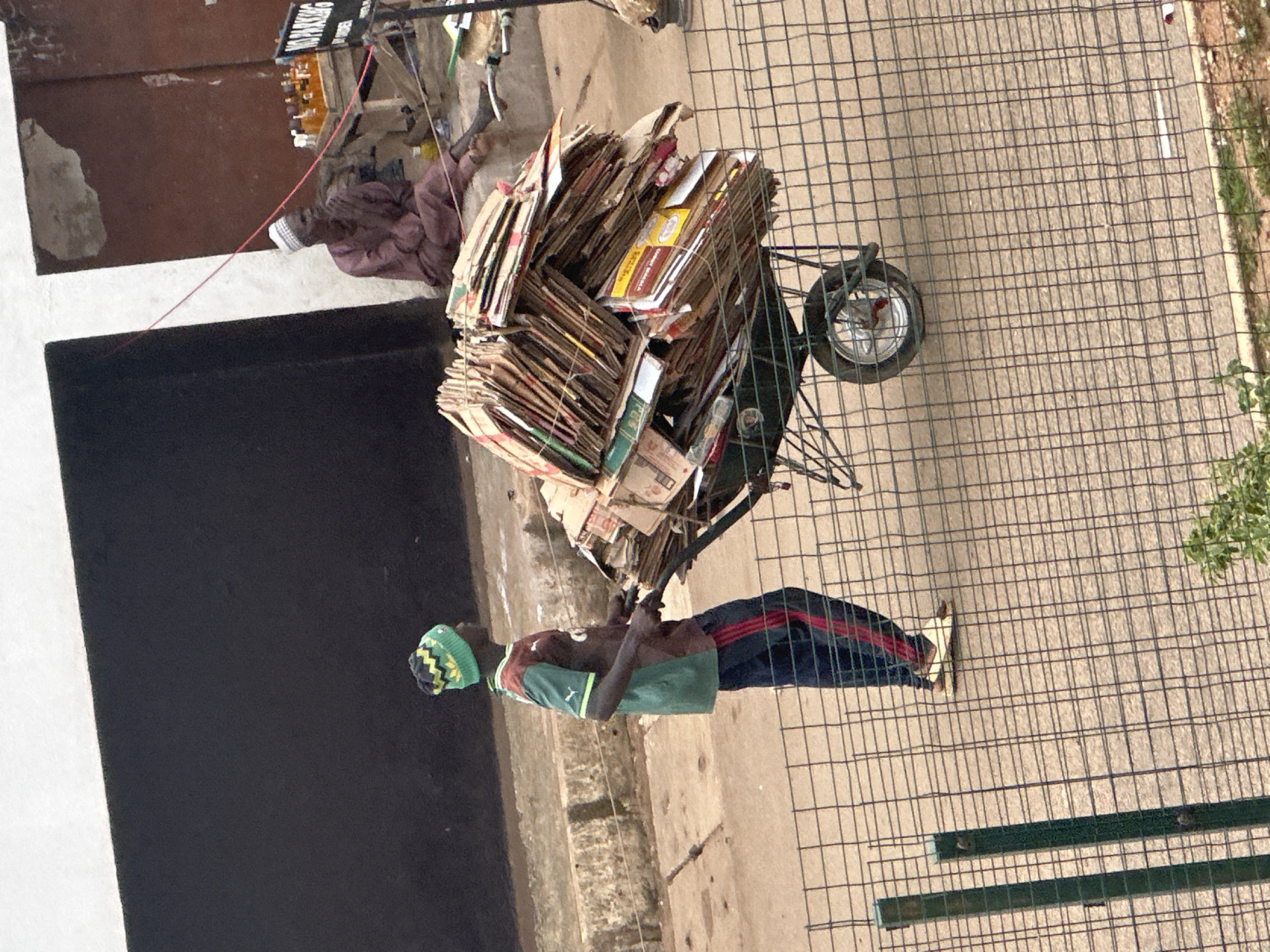 In Bukuru, Nigeria, a young man wearing a green and yellow beanie, a green and brown soccer-style jersey, and dark pants with red stripes is pushing a wheelbarrow loaded with a large quantity of bundled cardboard boxes. The wheelbarrow has one wheel and is a dark green color. In the background, an older man in a maroon garment and a white cap sits with his hands clasped, looking to the side. Behind him, there is a table with several bottles of amber liquid. A "NO PARKING" sign is also visible. The scene takes place outdoors, on a paved surface with a wire fence in the foreground. The overall impression is one of daily commerce and activity in a Nigerian urban setting.