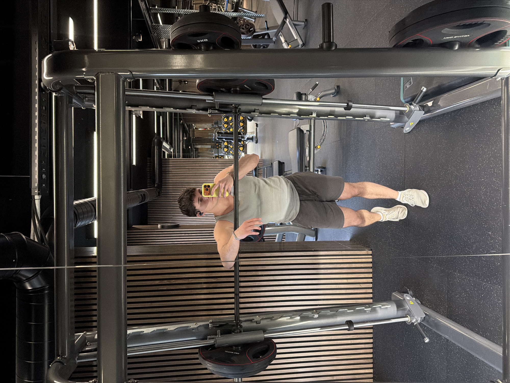 A mirror reflects a man in a gym in Sofia, Bulgaria. He is holding a yellow cell phone and wearing a light gray tank top, dark gray shorts, and white sneakers. The gym has black rubber flooring and a wall of horizontal wood slats. There is a Smith machine in the foreground with weight plates.
