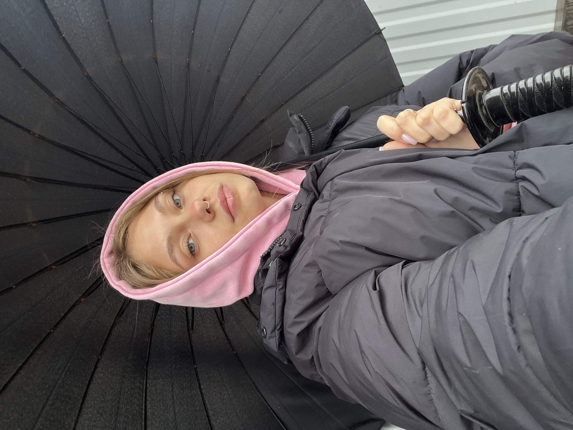 A young woman with blonde hair, wearing a pink hoodie under a black puffer jacket, is holding a closed black umbrella. She has piercings in her eyebrow and nose. The background appears to be an industrial building with a corrugated metal wall. The location is Фоки, Russia.