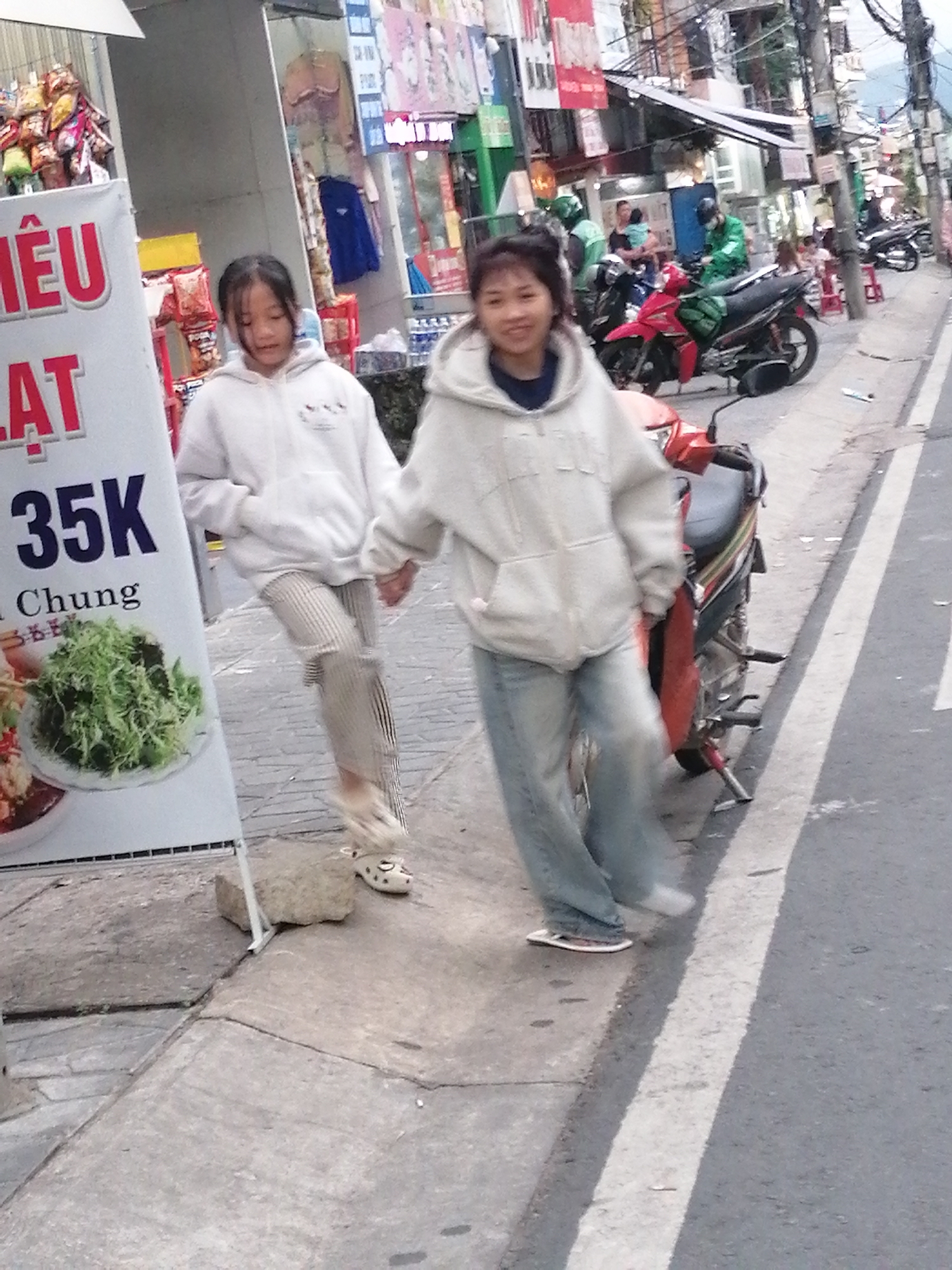 The image captures a street scene in Da Lat, Vietnam, with a focus on two young females walking on a sidewalk adjacent to a road. The sidewalk is paved with concrete and features a white line marking its edge. In the foreground, a white sign with red and black text, possibly advertising food or prices, stands prominently on the left. The sign has an image of a leafy green dish on it.
The two individuals are walking away from the camera, holding hands. The girl on the left is wearing a white hooded sweatshirt and striped pants, while the girl on the right is in a similar white hooded sweatshirt and blue jeans. Both are wearing casual footwear.
Behind the sidewalk, the street is visible, with several motorcycles parked or being ridden. Some individuals in green vests, likely delivery drivers, are present near some of the motorcycles. Buildings with various signs and awnings line the street, indicating a commercial area. Overhead, a complex network of electrical wires is apparent. The overall atmosphere suggests a busy urban environment.