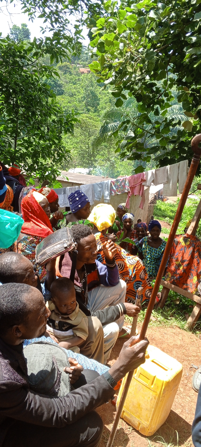 A group of people, mostly women, are gathered outdoors. Some wear colorful head coverings. A man holds a baby while seated. Another man stands nearby with a walking stick and a yellow jerrycan. Clothes are hanging on a clothesline behind the group. Trees and foliage surround the scene, with a glimpse of a valley in the background. The setting appears to be a rural or village environment.