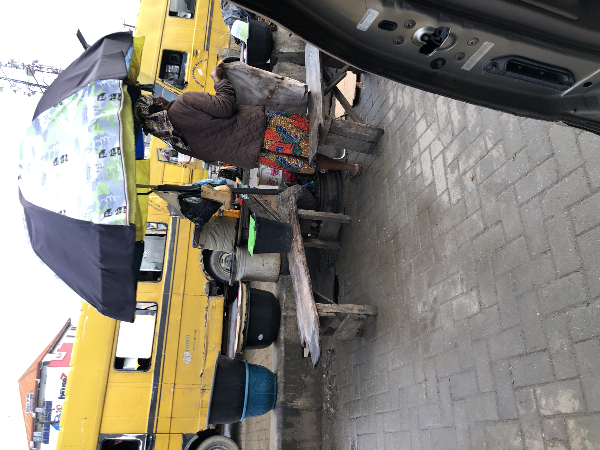In Ikeja, Nigeria, a street vendor operates under a large, colorful umbrella near a bright yellow bus. The bus, which appears to be a public transport vehicle, is parked on the side of a wet, paved road. In front of the bus, a woman dressed in a brown jacket and a vibrantly patterned skirt stands behind a wooden bench. She is seen holding a light-colored piece of fabric. Various containers, including black and blue buckets, are placed on a concrete barrier. In the background, buildings and communication towers are visible. The scene captures a slice of everyday urban life in Ikeja.