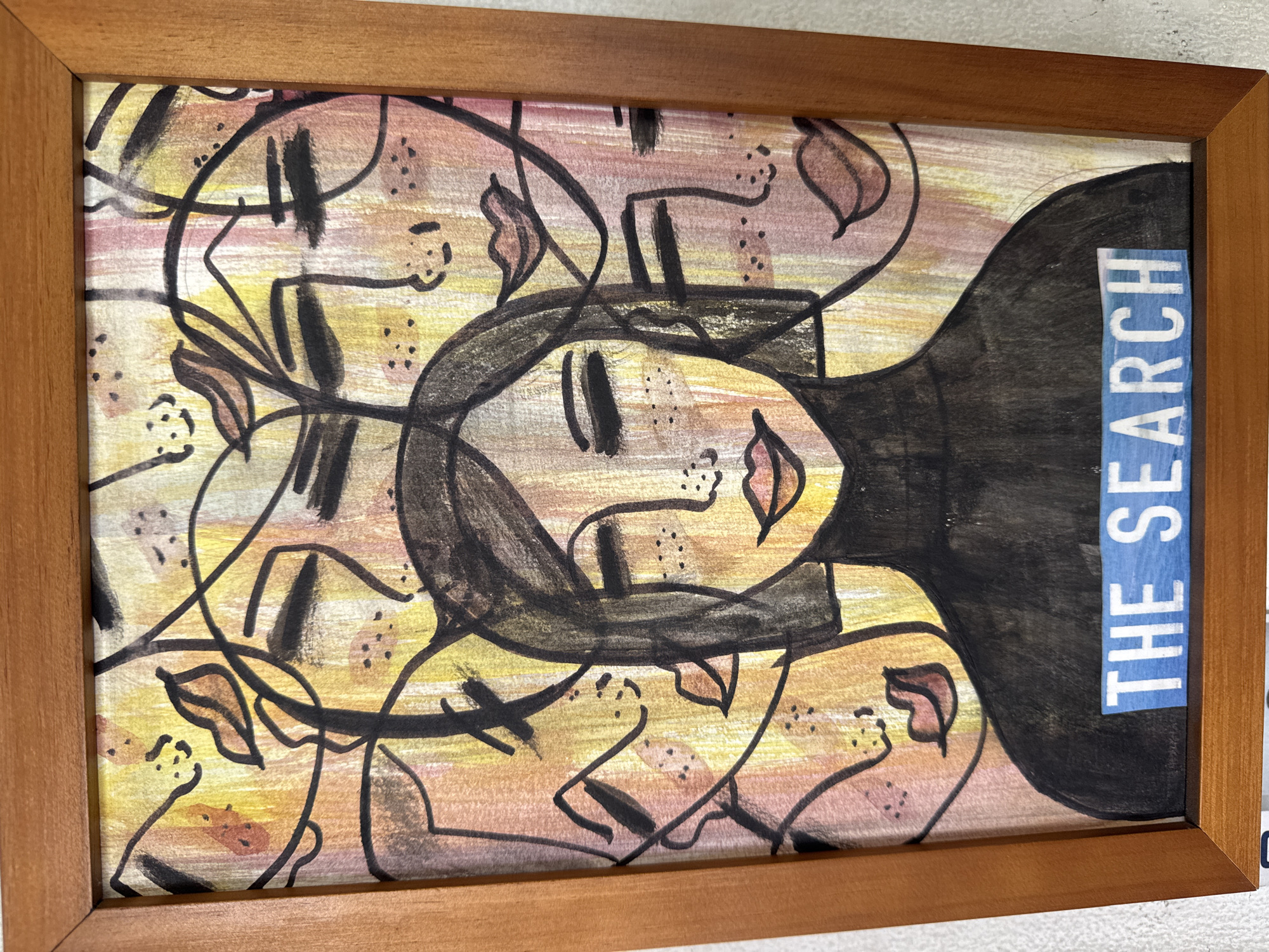 A framed abstract artwork titled "The Search" hangs on a light gray wall. The frame is made of brown wood. The artwork features a woman's face with elongated features and black hair. The words "THE SEARCH" are printed vertically on a blue and white background, positioned on the right side of the artwork, alongside the woman's hair. Abstract lines and other faces are drawn in the background, with a color palette of yellow, pink, and brown. The artwork is located in North Kuta, Indonesia.