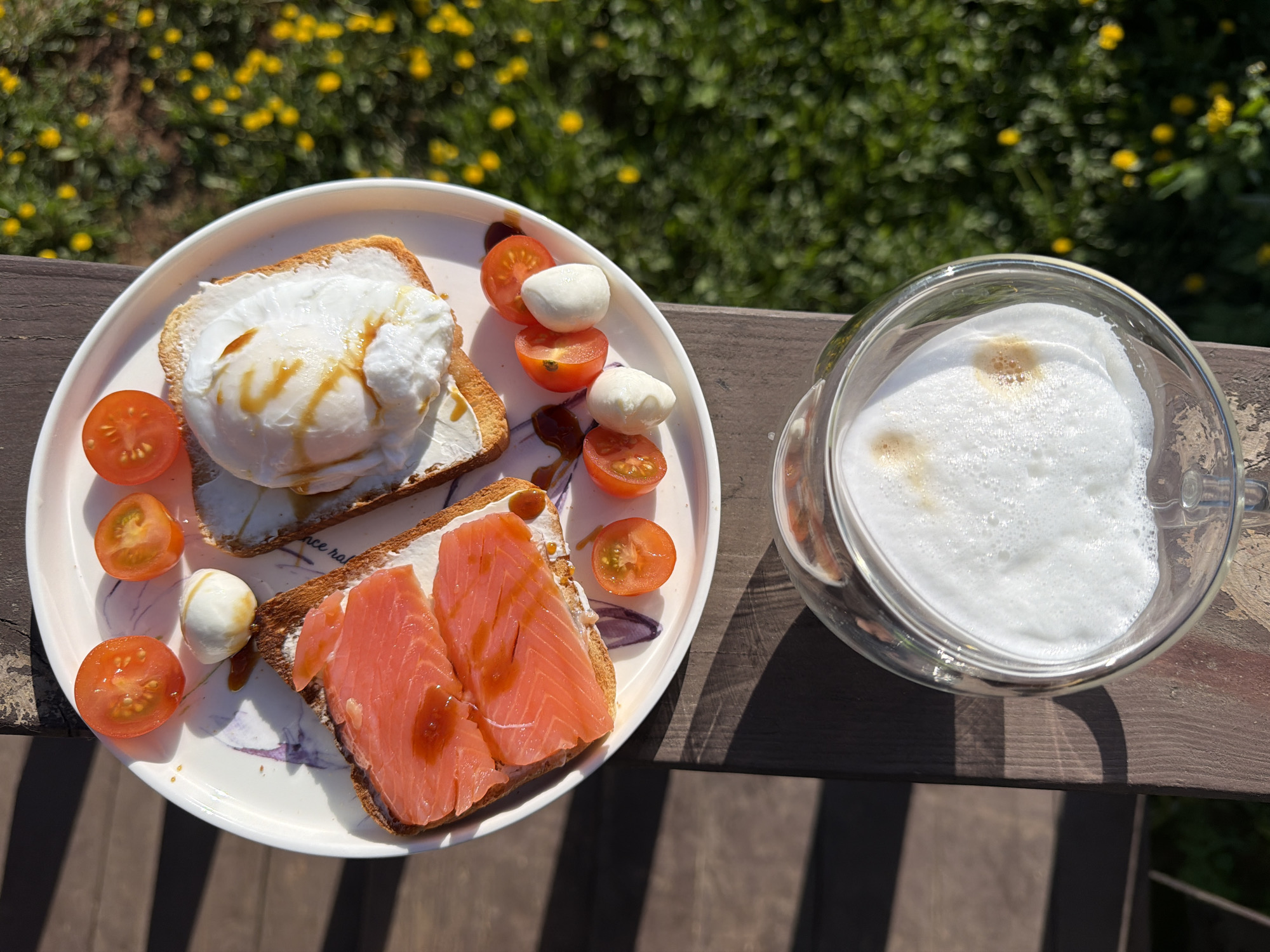 The image shows a plate with two slices of toast, each topped with different ingredients, alongside a glass of foamy beverage. The plate is placed on a wooden surface, possibly a table or railing, with greenery and yellow flowers visible in the blurred background.
One slice of toast is topped with a white spread, possibly cream cheese, and a poached egg drizzled with a dark sauce. The other slice of toast is also topped with a white spread and two slices of salmon, also drizzled with the same dark sauce. Sliced cherry tomatoes and small mozzarella balls are arranged on the plate around the toast.
The glass contains a white, foamy beverage with some brown spots, likely a cappuccino or latte.
It is not possible to confirm the location in the city of Chaikovsky, Russia, solely based on the visual information in the image.