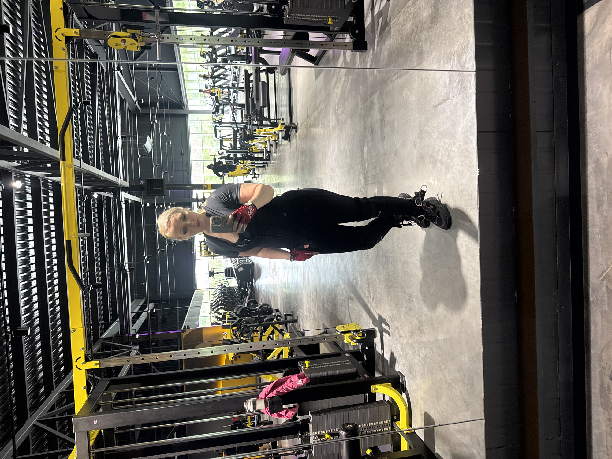 A woman is taking a mirror selfie in a gym in Komsomolsk-on-Amur, Russia. She is wearing black athletic clothing, red and black gloves, and black athletic shoes with pink accents. The gym features yellow and black weight machines, a concrete floor, and a high ceiling with industrial lighting and exposed pipes. Other gym-goers are visible in the background using treadmills. The overall environment suggests a modern and well-equipped fitness facility.