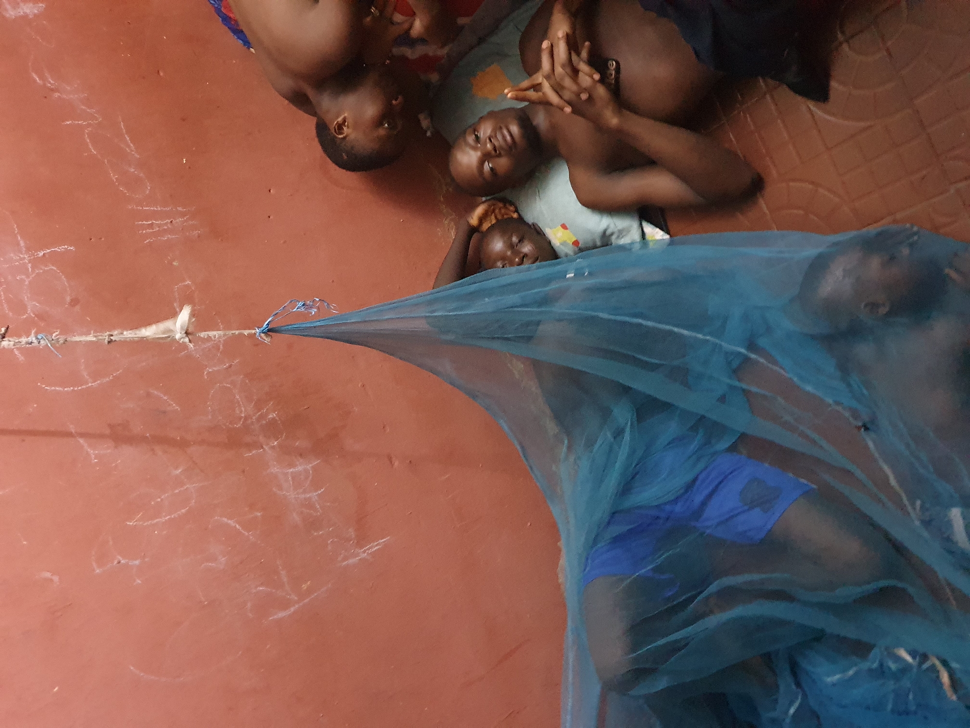 The image captures a group of individuals, appearing to be children or young adults, resting on a reddish-brown floor indoors. At least five individuals are visible, with three clearly in the upper left section, and two more partially obscured by a blue mosquito net in the lower right.
In the upper part of the frame, three individuals lie close together on a light-colored mat or blanket. Their faces are calm, and they appear to be either sleeping or resting. One individual in the center has their hands clasped. A fourth individual's head is partially visible in the top left corner.
The lower right section of the image is dominated by a bright blue mosquito net, stretched diagonally across the frame and tied to a point on a wall or post on the left. Beneath the net, the body of an individual, wearing blue shorts, is visible, suggesting they are also resting. The use of a mosquito net indicates a measure against mosquito-borne diseases, common in regions like Idah, Nigeria.
The floor is predominantly a reddish-brown hue, possibly concrete or a similar material. In the top right corner, a section of the floor appears to be tiled with a brown patterned design. Scattered across the reddish-brown floor are numerous abstract white chalk markings or scribbles. These markings do not form legible words or distinct images but rather appear as random doodles. The lighting is subdued, suggesting an indoor setting perhaps during evening hours or a relaxed moment of the day. The overall atmosphere conveyed is one of quiet rest.