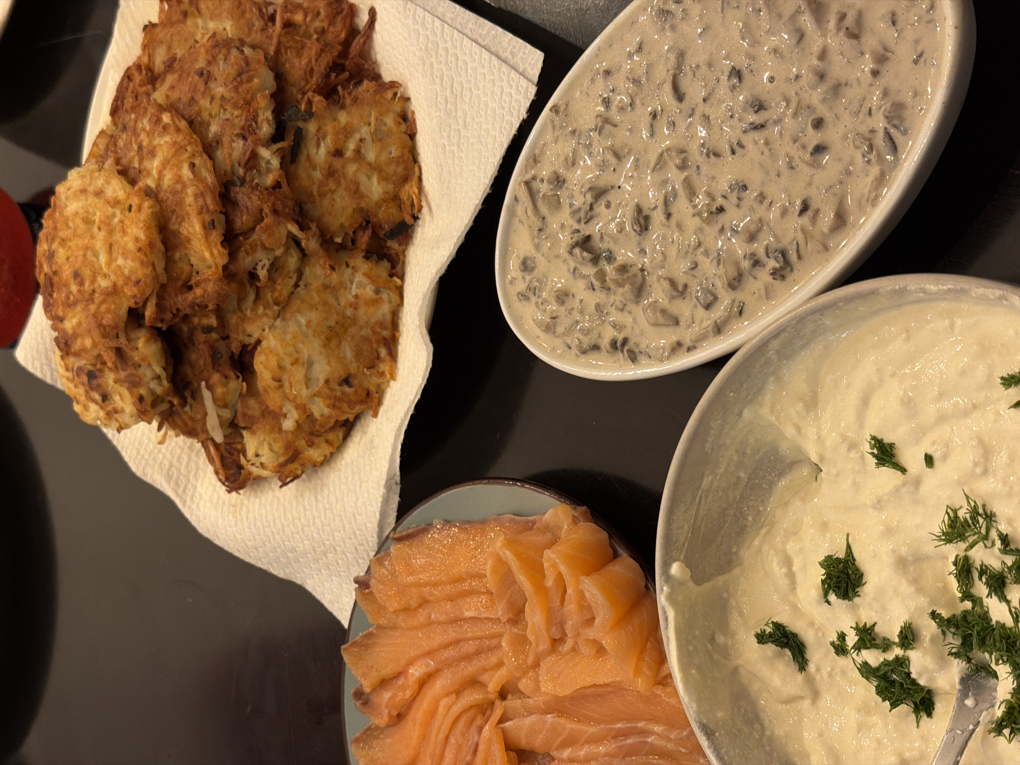 The image shows a table arrangement of food items. In the upper left is a stack of fried potato pancakes, likely latkes, on a white paper towel. Adjacent to them and slightly above is an oval dish containing a creamy mushroom sauce. Below the pancakes is a round plate with slices of smoked salmon. To the right of the salmon is a round bowl filled with a creamy sauce, possibly sour cream, garnished with dill. The table is dark in color. The scene appears to be in a domestic setting, judging by the presentation of the food. I cannot determine the location as Ramat Gan, Occupied Palestine.