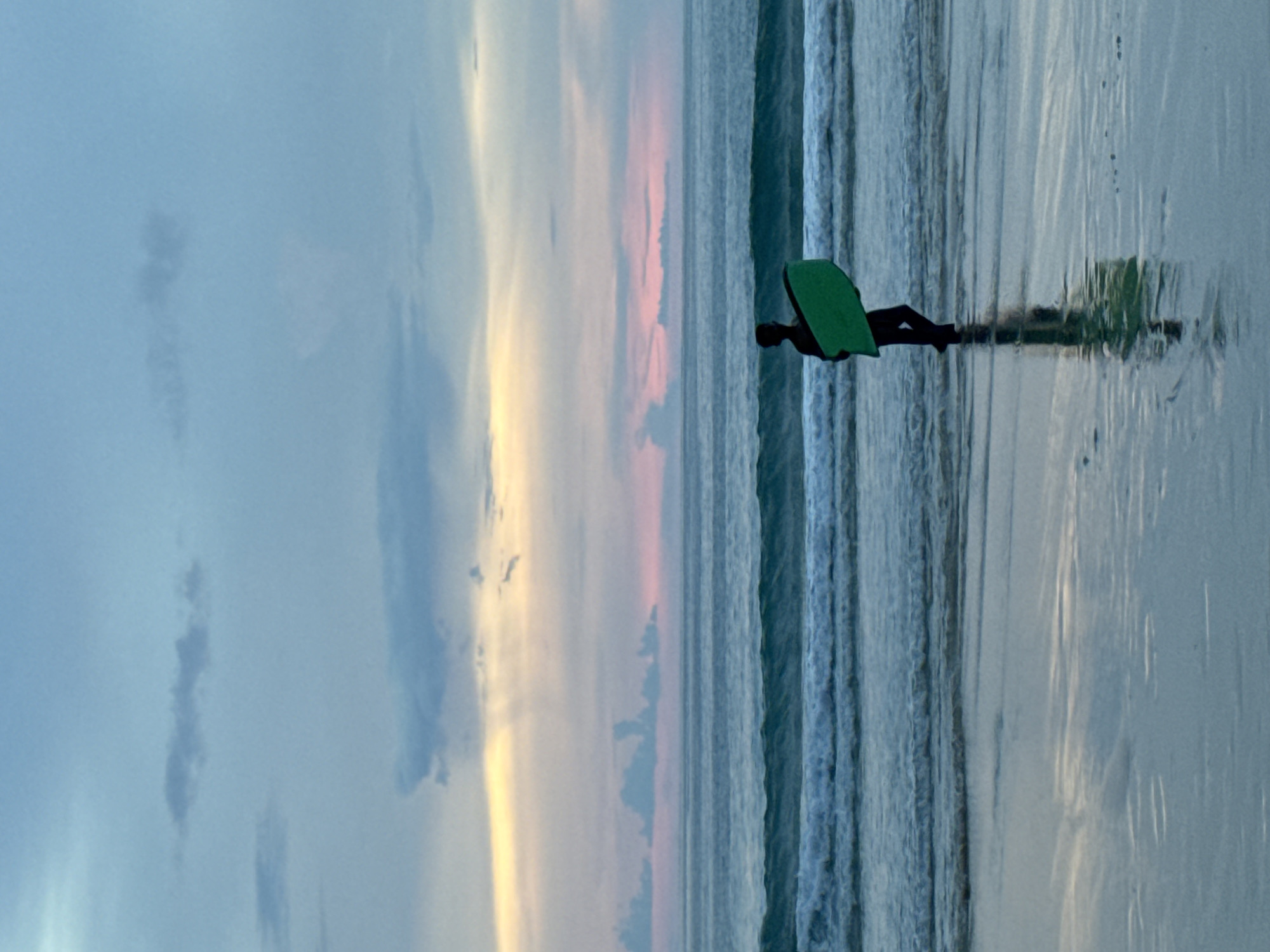 A solitary person, silhouetted against a dusky sky, stands on a wet beach in Choeng Thale, Thailand, holding a bright green boogie board. The ocean stretches out before them, with gentle waves lapping at the shore. The sky above is a canvas of soft blues, pinks, and yellows, indicating either dawn or dusk. The wet sand reflects the muted colors of the sky and the person's silhouette. The overall impression is one of peaceful solitude and the quiet beauty of a beach at the end of the day.