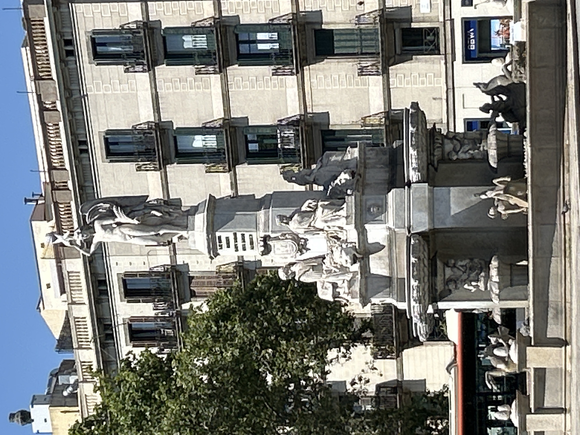 This is a medium shot of a large, multi-tiered stone fountain in Barcelona, Spain, on a sunny day. The fountain features intricate sculptures of figures and mythical creatures, with water cascading down. Behind the fountain, a large, light-colored building with multiple floors and balconies can be seen. The building has rows of dark-colored windows and balconies, some of which are adorned with decorative railings. A large, leafy green tree is partially visible to the left of the fountain, obscuring some of the building. A red and silver bus is visible in the background, to the left of the fountain. The sky is a clear blue. The overall scene depicts a public square with a prominent artistic monument.