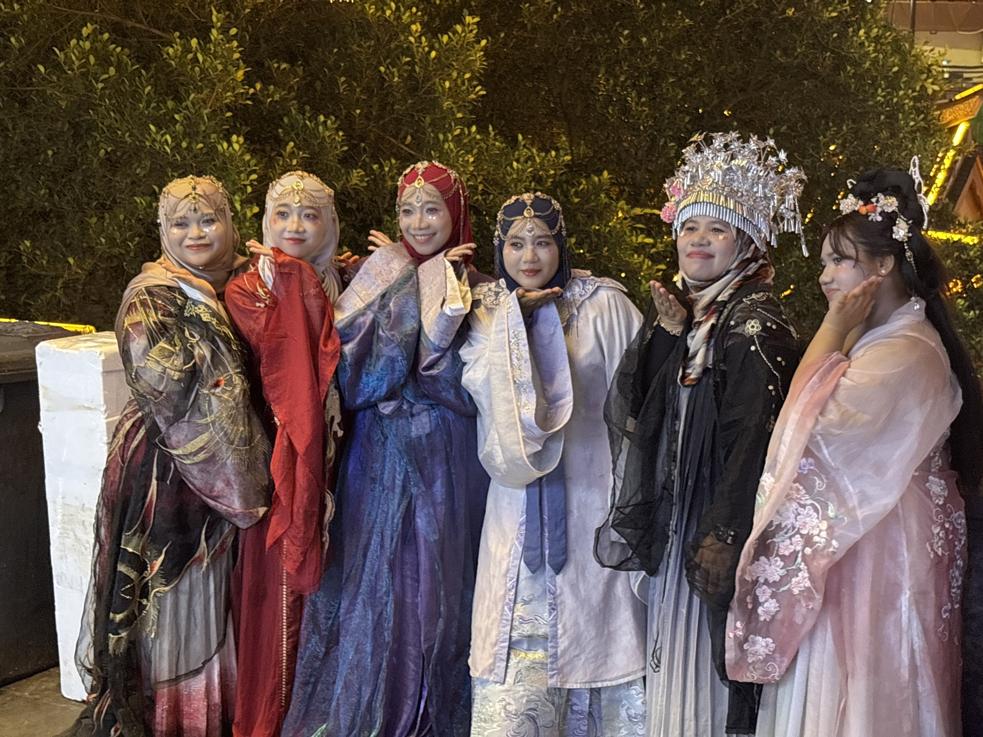 Six individuals are aligned in a row, standing in front of a backdrop of dense foliage, likely in an urban area. They are all dressed in elaborate, traditional Chinese attire. Each is wearing a head covering of varying design. They are posing with their hands close to their faces, creating a similar hand gesture among them. Their facial expressions are cheerful. The location, based on the metadata, is likely Yuzhong District, China.