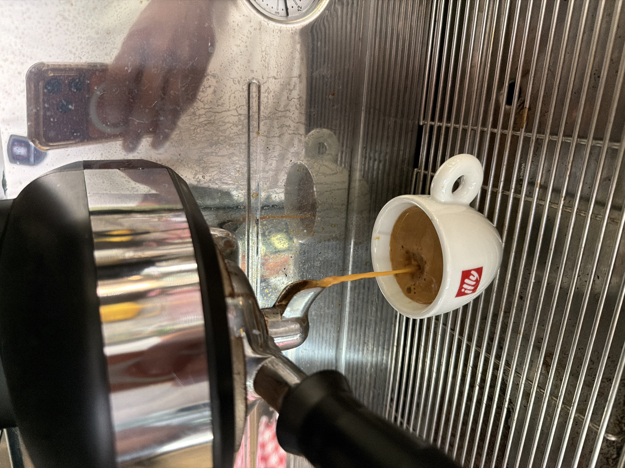 An espresso is being brewed into a white Illy cup. The espresso machine is stainless steel with a black top. A hand holding a smartphone is reflected in the machine. The scene is inside a cafe in Sofia, Bulgaria.