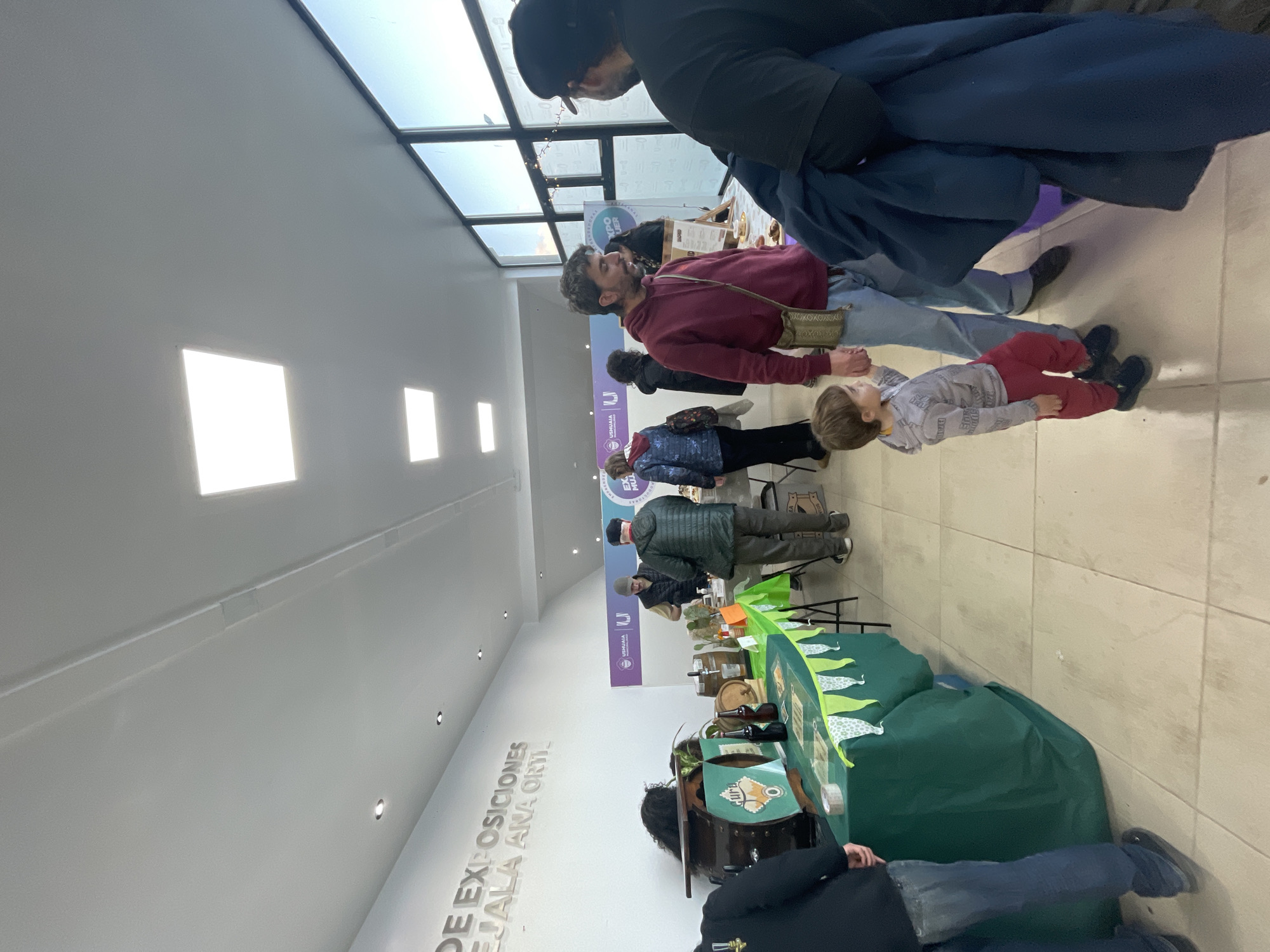 The image depicts an indoor scene, likely a market or exhibition space in Ushuaia, Argentina. A group of people are present, including men, women, and a child. One man is holding the hand of the child, who is wearing red pants and a patterned grey top. The man wears a red jacket and a crossbody bag. A person wearing a black hat and black coat stands near them.
Several booths are set up, covered with green tablecloths and decorated with animal-themed designs. The inscription "DE EXPOSICIONES" and what appears to be "USHUAIA" can be seen on a white wall behind the booths.
The location is brightly lit by overhead skylights and square ceiling lights. The floor is tiled. The overall impression is that of a local community event.