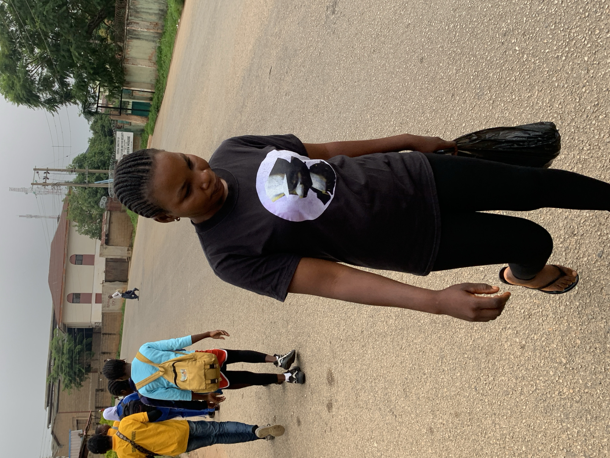 A group of people walk on a paved road in Jos, Nigeria. In the foreground, a young woman with braided hair, wearing a black t-shirt with a circular graphic and black leggings, walks away from the camera, carrying a black plastic bag in her right hand. Behind her, other individuals are walking in the same direction. One person wears a bright yellow t-shirt, another a blue shirt with a yellow backpack, and a third person wears a blue shirt and a white headscarf. The road is lined with buildings and trees, and power lines are visible in the distance. The sky is overcast.
