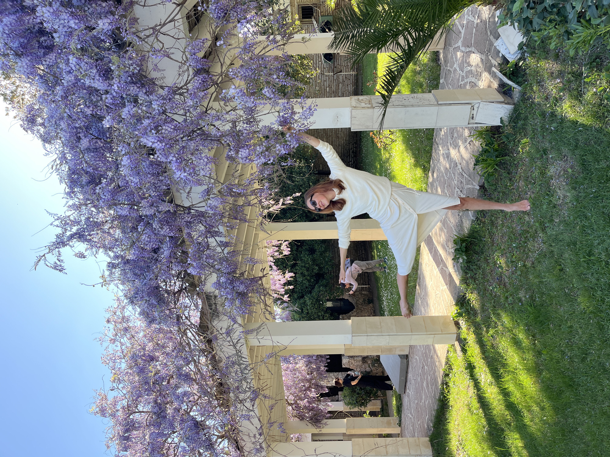 A woman wearing sunglasses and a white outfit stands on a paved walkway under a pergola covered in purple wisteria flowers. Her left arm is extended towards the flowers, and she balances on one leg. Behind her, other individuals are partially visible between the pergola supports. The area features green grass, a palm tree, and stone structures. The location is Boreti, Montenegro.