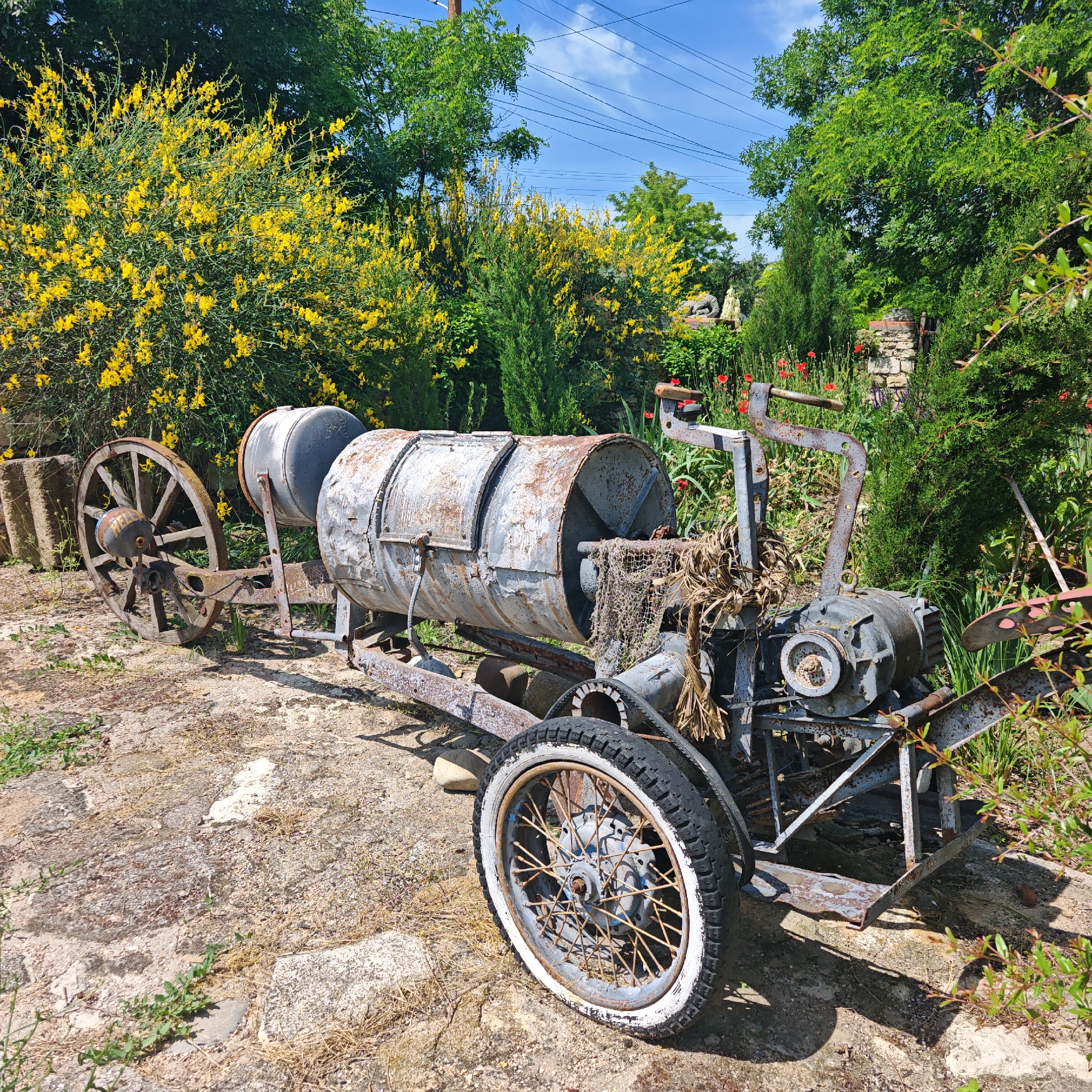 The image depicts an outdoor scene in Останинское сельское поселение, Ukraine, featuring a heavily rusted, antiquated piece of machinery positioned on an uneven dirt and concrete surface. The central object consists of a metal frame supporting two large, horizontally oriented cylindrical drums. The drum on the left is smaller and smoother, while the larger drum on the right exhibits visible seams, a side access panel, and internal baffles visible through an end opening.
The machinery is equipped with two dissimilar wheels. On the left, a large wooden spoked wheel with a metal hub, marked "MIL," is attached. On the right, a smaller, spoked wheel with a worn white-walled tire, characteristic of a motorcycle or vintage bicycle, is present. An exposed motor or engine component with a pulley and belt system is visible on the right side of the machine, near the smaller wheel. Ropes and a segment of netting are draped over various parts of the rusted structure.
The environment is characterized by abundant vegetation under a clear blue sky with sparse white clouds. Prominently in the background and to the left, dense bushes covered in small yellow flowers (likely Spanish Broom) are visible. Darker green trees are situated further back. Near the right side of the machinery, patches of red poppies are interspersed among other green foliage. A wooden utility pole with multiple overhead power lines is visible against the sky in the upper left. A low stone wall with a flat cap is situated in the middle-right background. The ground beneath the machinery is a mix of bare earth, dried grass, scattered stones, and segments of concrete paving. Bright sunlight casts distinct shadows across the scene. No individuals are present, and no actions are being performed.