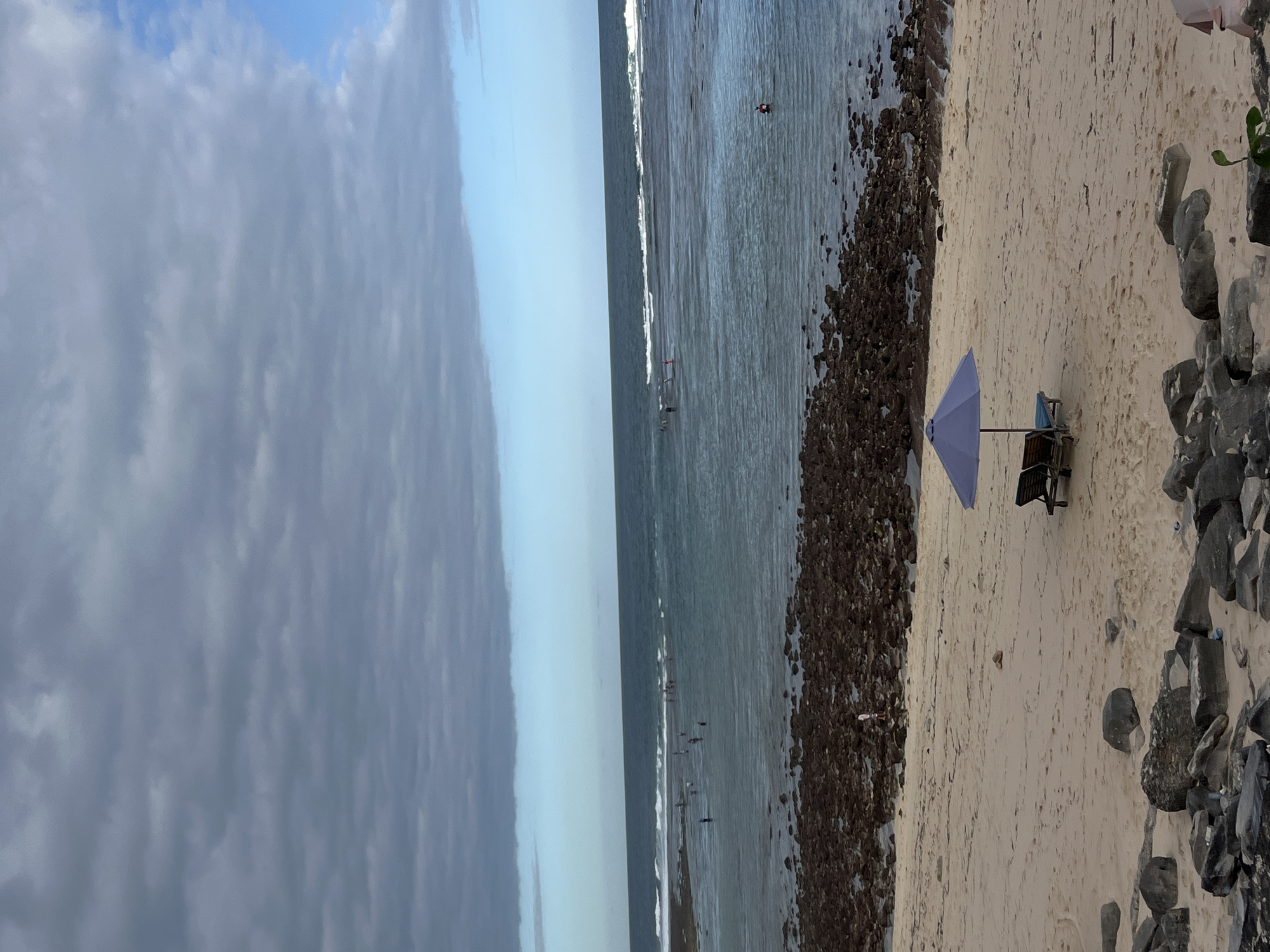This photograph depicts a beach scene in Kuta Selatan, Indonesia. The sky is a gradient of gray clouds transitioning to light blue towards the horizon. The ocean is visible with small waves and several people are in the water. A dark rocky area separates the water from the sandy beach. A light purple umbrella and a chair are on the beach, which is flanked on the landward side by darker rocks and some vegetation.