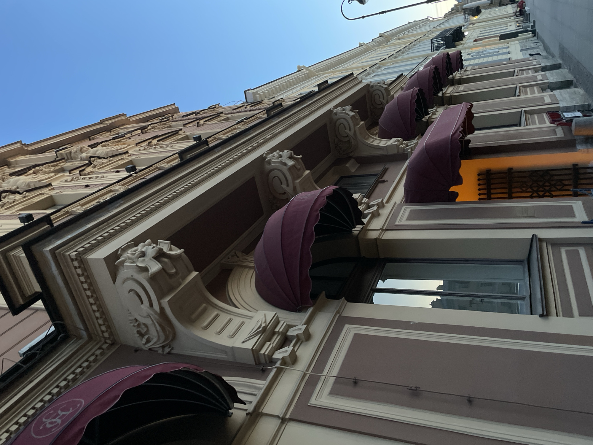 The photograph presents a dynamic, low-angle, and tilted view of an ornate, multi-story building facade in Saint Petersburg, Russia, set against a clear blue daytime sky. The architecture is distinctly classical, characterized by intricate detailing including decorative pilasters, cornices, elaborate corbels with sculptural elements, and various moldings, rendered in a palette of cream, beige, and brown.
Several retractable awnings, in a rich maroon or deep purple hue, are partially unfurled over windows on the lower levels. One awning notably features a white, stylized logo, which appears to be "S&S" or "CS". The windows reflect portions of the sky and adjacent structures. Near the street level, a window emits a warm, orange glow, suggesting an active shop or café, protected by a decorative black metal grate. A curved black streetlight fixture is visible in the upper right against the bright sky. The clear weather and absence of visible people allow the focus to remain on the building's historical grandeur and detailed craftsmanship.