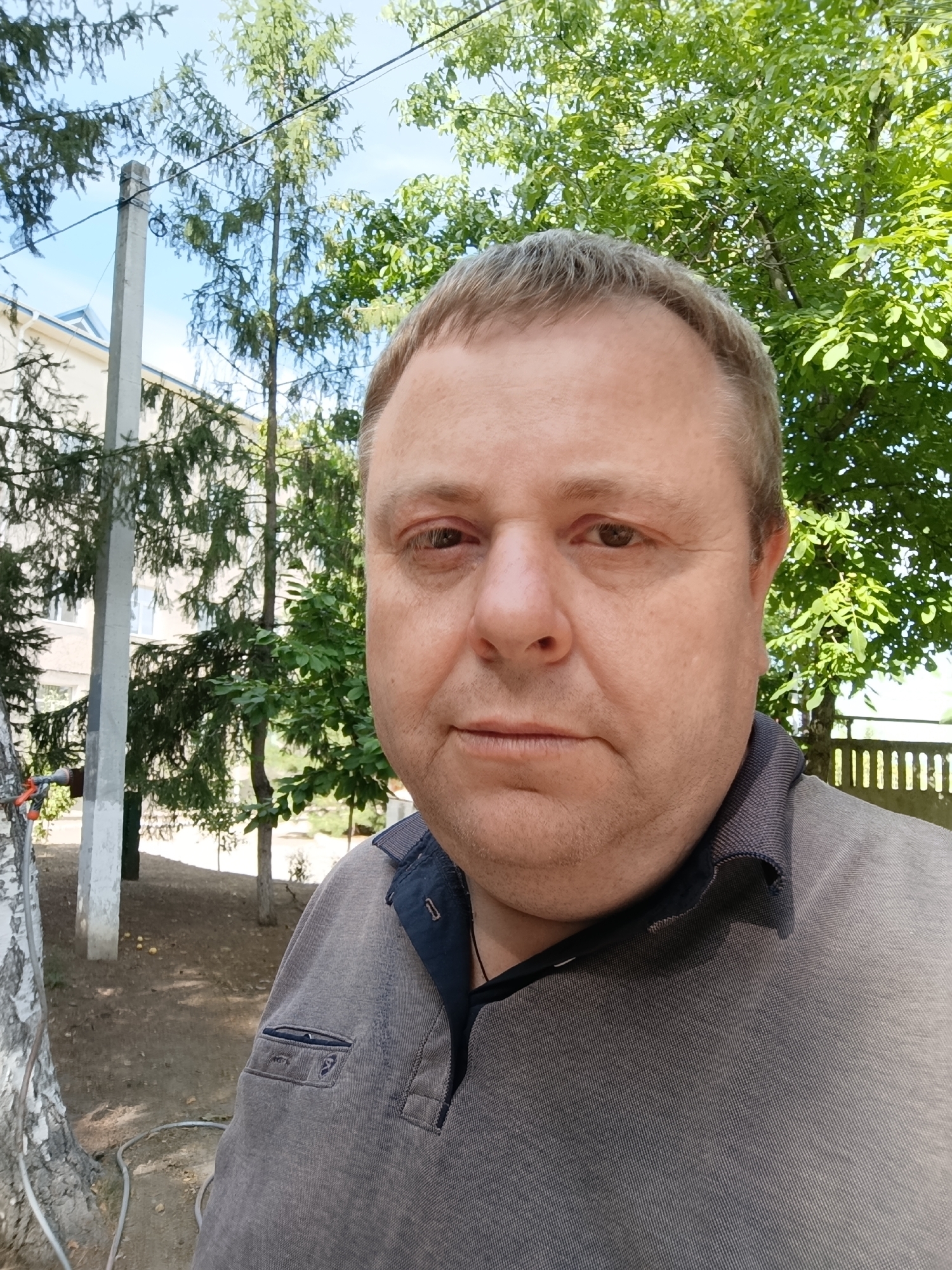 A mid-shot, selfie-style portrait captures a middle-aged man with fair skin, light brown hair, and light brown eyes. He is looking directly at the camera with a neutral expression. His left eye appears slightly red. He is wearing a brown collared polo shirt with a dark blue inner collar and trim.
The background suggests an outdoor setting, likely in a town or residential area. Behind the man, a large tree with vibrant green leaves dominates the right side of the frame. To the left, a light-colored building with a blue-tiled roof is partially visible. A concrete utility pole stands near the building, and a white-barked tree is on the far left. The ground appears to be covered in dirt and fallen leaves, with some small yellow objects scattered on it. A thin, dark fence is visible behind the man to the right. The overall lighting indicates it is daytime, with clear, bright sunlight.
Given the context of the request, it is plausible that this image was taken in Dubăsari town council, Moldova, a region known for its rural and semi-urban landscapes with similar architectural styles and vegetation.