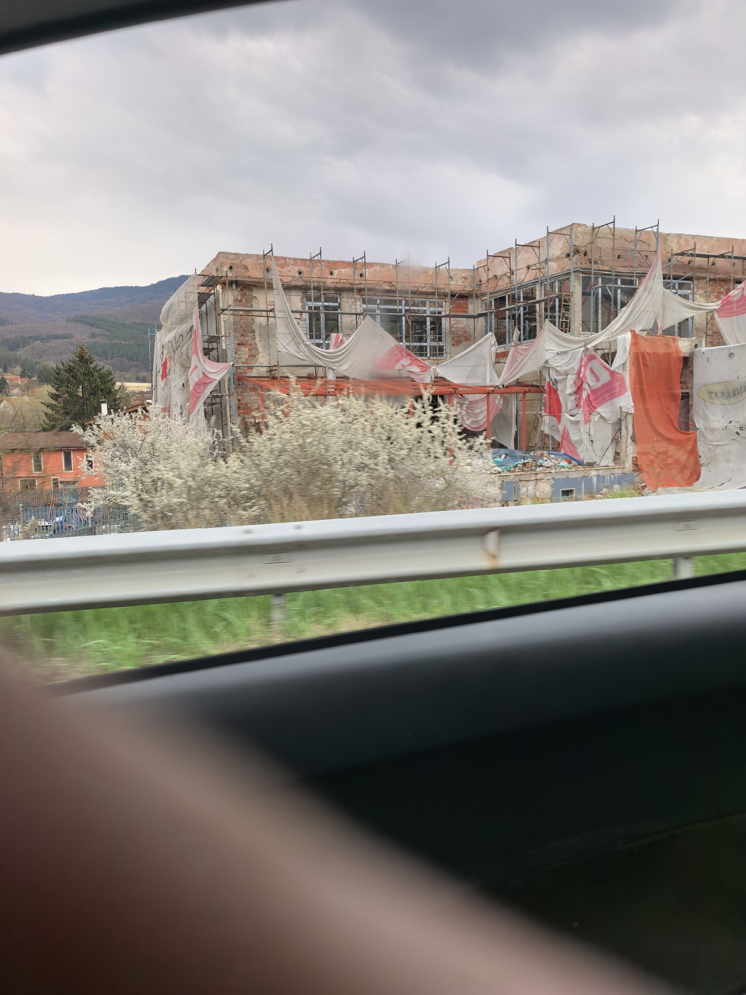 The media file captures an outdoor scene from the interior of a moving vehicle, evidenced by the blurred foreground and car parts visible at the bottom. The central subject is a large, multi-story building under construction or renovation, or possibly in a state of disrepair. It features exposed brickwork, peeling plaster, and numerous window openings, some fitted with frames and others empty.
Extensive metal scaffolding surrounds the building, covered in large white and orange tarpaulins or construction nets. These coverings are draped and secured, with some white sections bearing red patterns or logos. Visible text includes "ТЕРАФЛЕК" (TERAFLEK) in Cyrillillic script on a white tarp on the far right, and partially legible words like "VELCO" or "VCLCO" on other red and white coverings.
In front of the building, several white-blossoming trees or large bushes are in full bloom, suggesting a springtime setting. Beyond the construction, rolling hills or mountains covered with trees extend into the background. The sky is overcast and cloudy, diffusing the daylight.
A white metal road guardrail runs horizontally across the mid-ground, partially obscuring blurred green grass beneath it. The very bottom of the image shows the dark interior of the vehicle, with a blurred, fleshy object (possibly a finger or hand) visible in the lower-left corner. The scene depicts an observation of a building project or abandoned structure within a natural landscape.
**Location context: Unknown, Unknown**