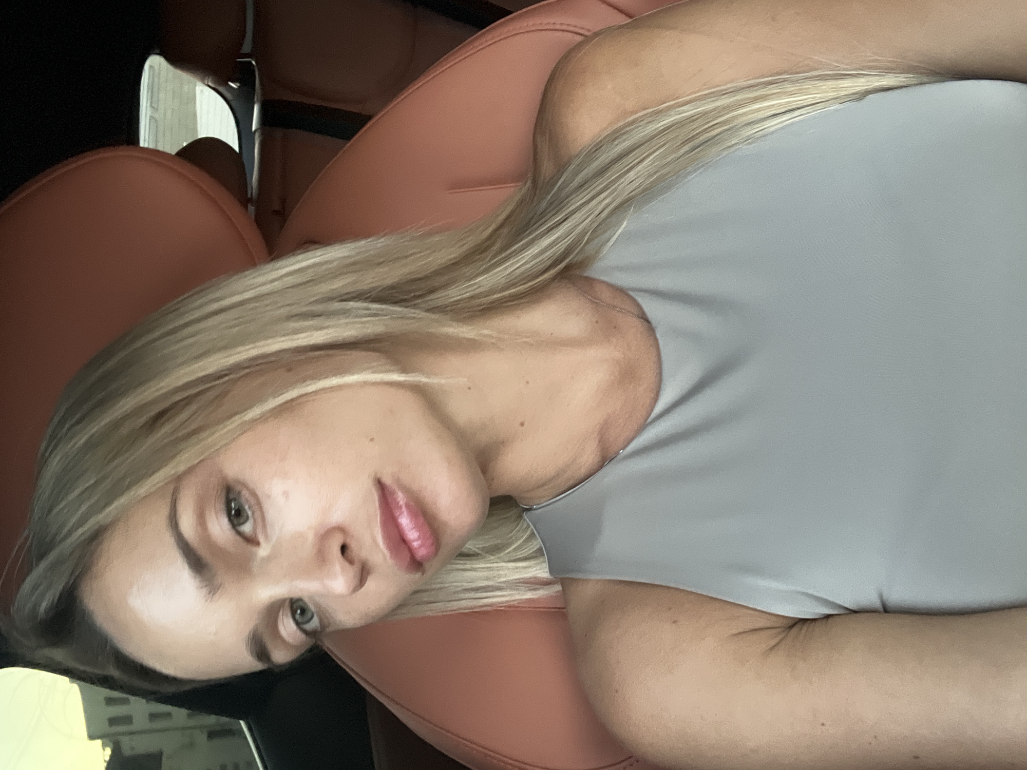 A woman with blonde hair and blue eyes is seated in a car. She is wearing a grey sleeveless top. The car has orange leather seats. The background shows buildings and a glimpse of the sky, suggesting an urban environment. The lighting suggests it is daytime, possibly late afternoon or early evening. The image appears to be a selfie taken by the woman. The location is stated to be Sharm El Sheikh, Egypt.