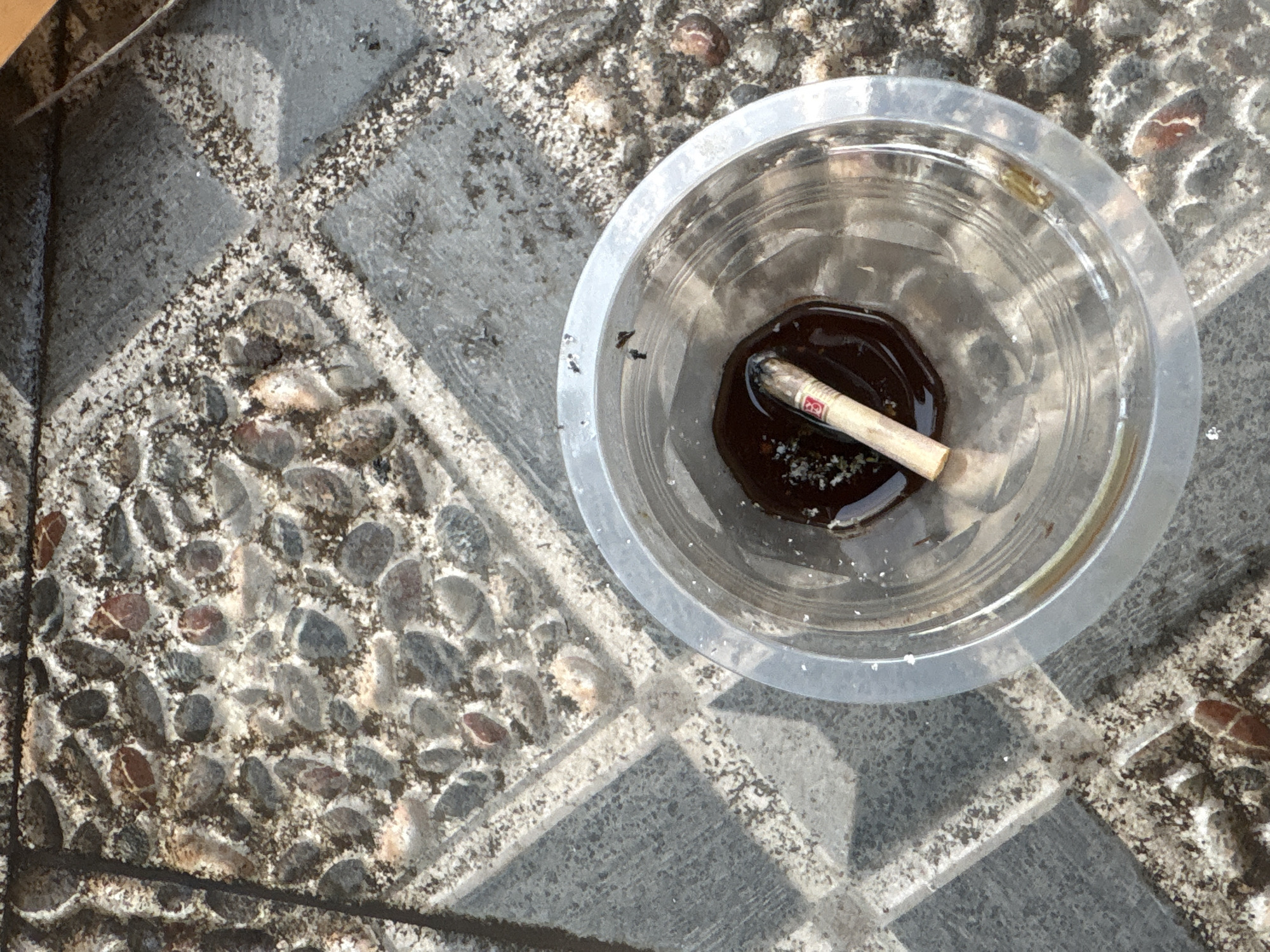 The image shows a close-up, overhead view of a clear plastic cup on an outdoor tiled surface. The cup contains dark liquid and a single cigarette butt, which appears to be extinguished. The surrounding tiles are made of concrete with embedded stones and have a pattern of diagonal lines. The lighting suggests it is daytime, with shadows cast across the tiles. The overall scene is one of neglect or litter, with the cigarette butt in the cup suggesting a casual disposal. The location is identified as Palimanan, Indonesia.