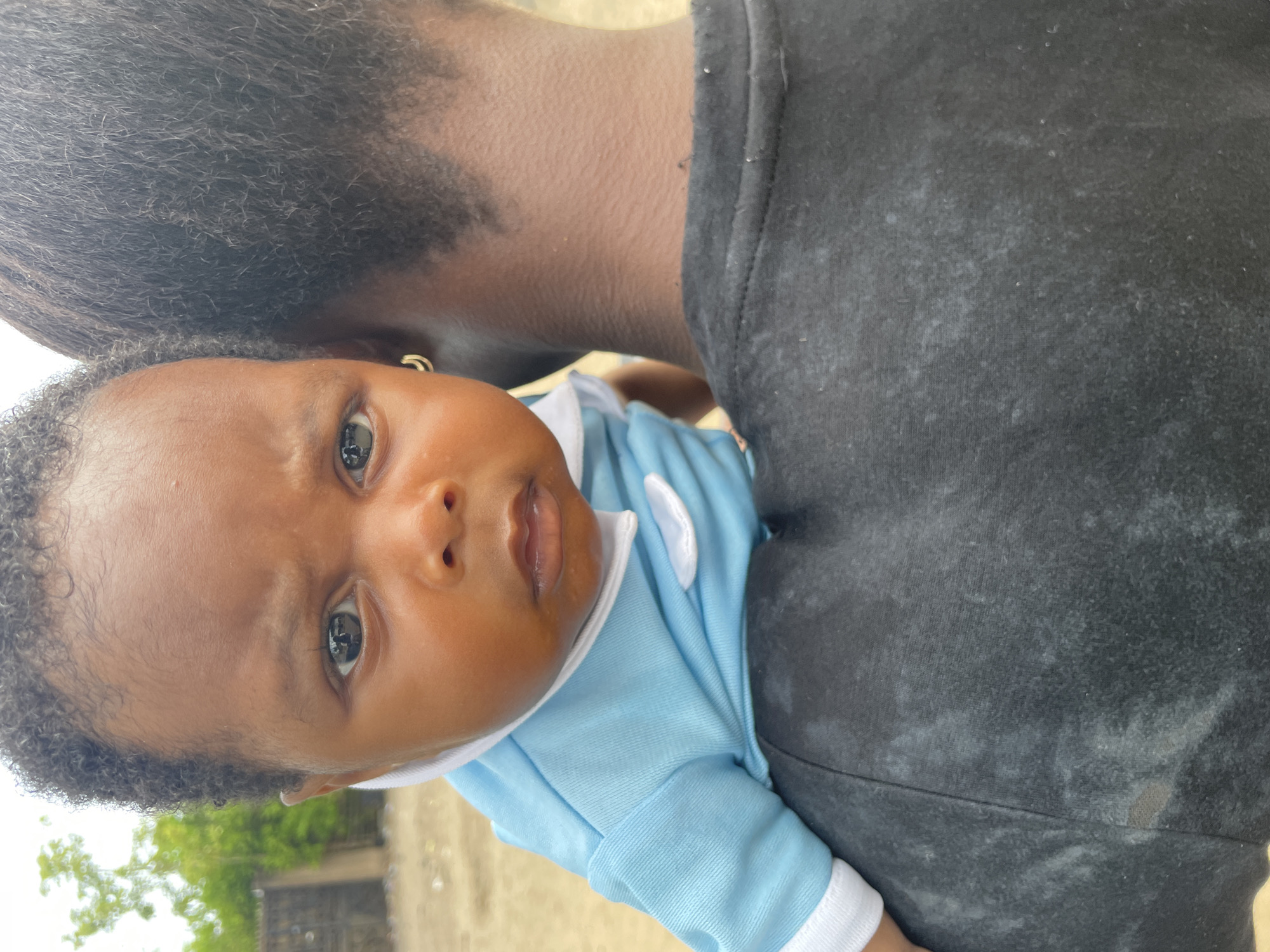 A baby is being held by a person with dark hair. The baby is wearing a blue onesie with white trim. The person holding the baby is wearing a black shirt. The scene is in daylight. The location is indicated as Badagry, Nigeria, suggesting an outdoor or possibly indoor setting.