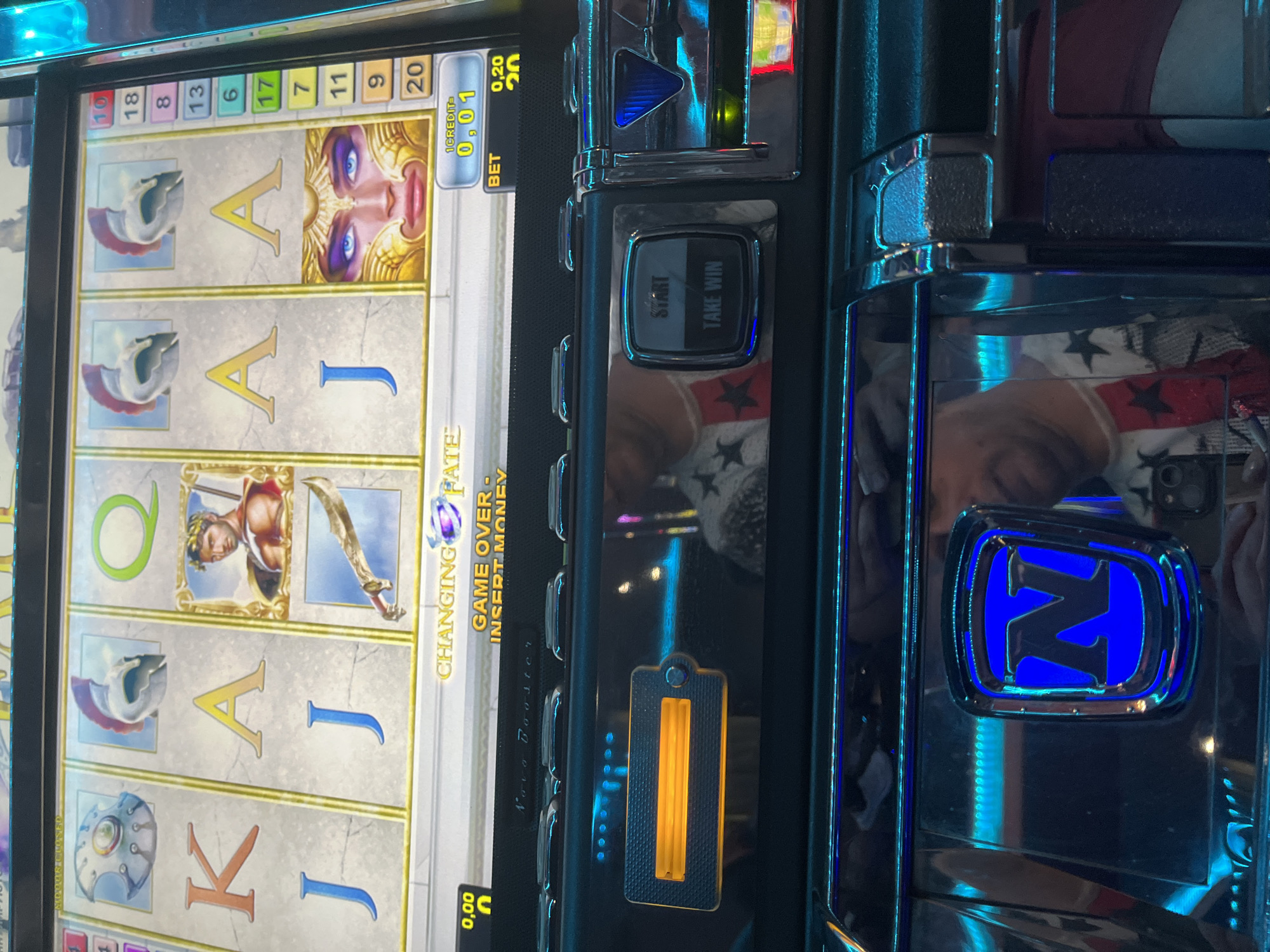 The image displays a close-up view of a casino slot machine in an unknown location. The machine's screen shows a game with an ancient Greco-Roman theme, featuring symbols such as a shield, two helmets, a male figure wearing a laurel wreath, a sword, and a female face in a golden headdress, along with the letters K, Q, A, and J. Textual elements on the screen include "CHANGING FATE", "GAME OVER - INSERT MONEY", "1 CREDIT=0,01", and "BET 0,20". A column of numbers ranging from 6 to 20 is present on the right side of the screen, likely indicating paylines. Below the screen is the machine's control panel, featuring a rectangular slot illuminated with an orange-yellow light, presumably for currency or credit card insertion. To its right, a square button is visible, labeled "START" and "TAKE WIN". The glossy surface of the machine's panel prominently reflects a person, visible from the torso up, wearing a white garment adorned with black and red star patterns, holding a dark-colored smartphone with a visible camera lens. The text "Novo Booster" is faintly discernible on the panel's upper surface. At the bottom of the machine, a blue illuminated "N" logo is displayed. The immediate environment beyond the machine is not discernible.