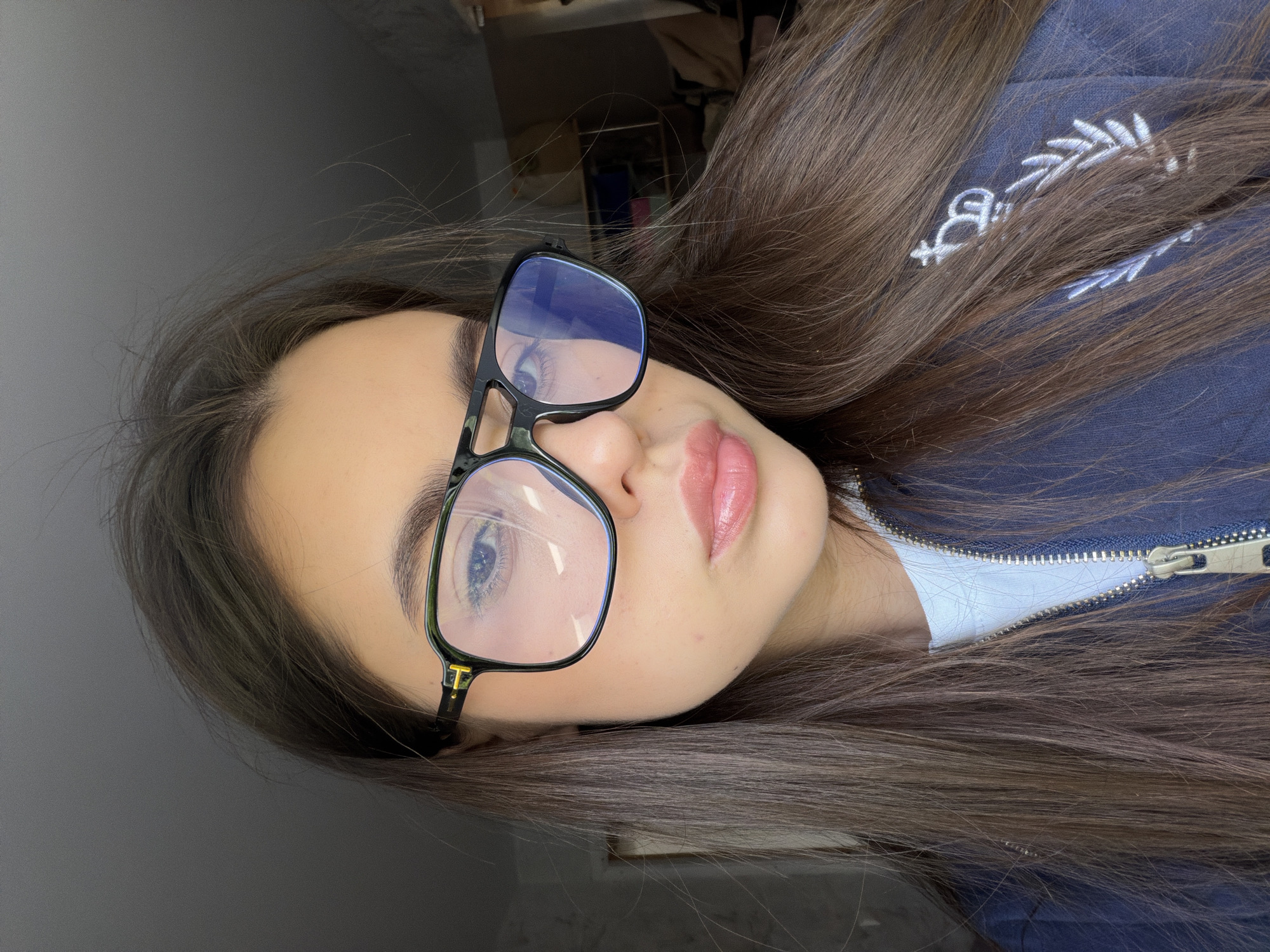 A woman with long brown hair is lying down, facing the camera. She is wearing glasses with black frames. Her lips are glossy. She is wearing a blue jacket with a zipper. A white shirt is visible underneath the jacket. The background is a plain gray wall. The image was not taken in Chaikovsky, Russia.