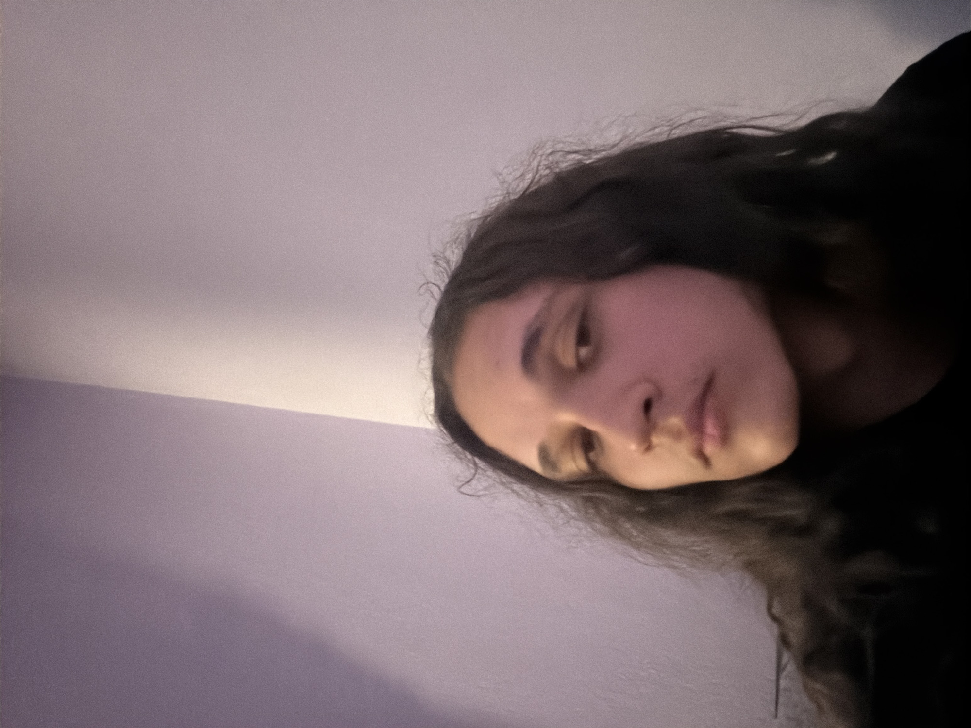 The image is a selfie-style, close-up shot of a person's face and upper body. The individual has long, dark, wavy hair. The person's face is softly lit, showing details of their skin and facial features. The background is a pale purple, possibly a wall. The lighting in the room seems somewhat dim. There is no discernible location or information related to Asenovgrad, Bulgaria.