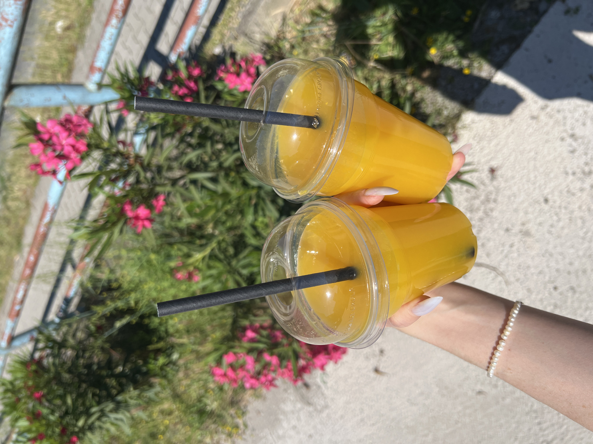 A person holds two clear plastic cups filled with an orange liquid, likely juice or a smoothie. Each cup has a black straw inserted through the lid. The background shows a glimpse of a weathered blue railing, pink oleander flowers, and greenery. The scene appears to be outdoors. The person is wearing a pearl bracelet on their wrist. The location is unknown.