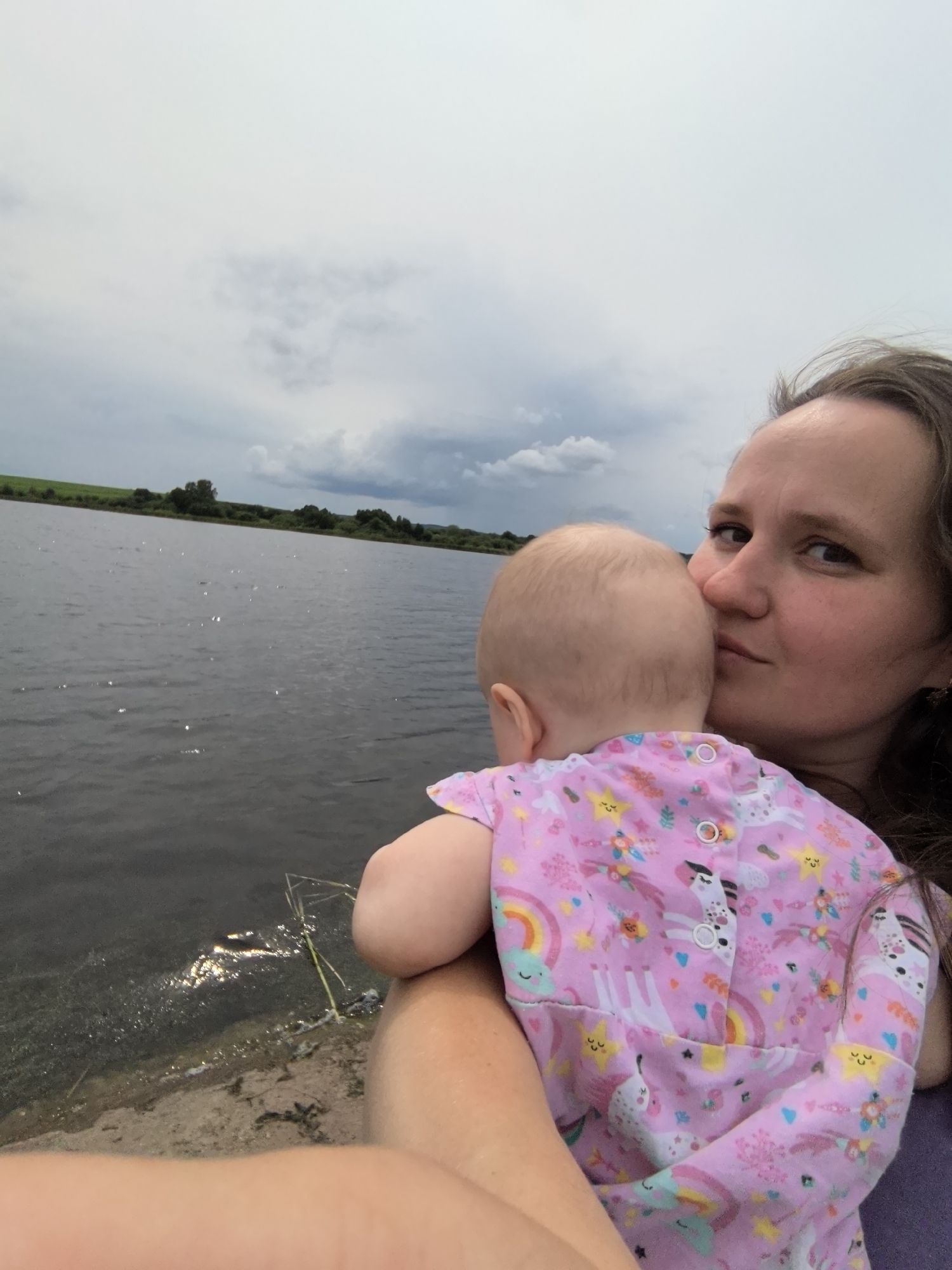 A woman is holding a baby near a body of water. The woman is on the right side of the image, looking towards the camera with a slight smile. The baby is facing away from the camera, nestled against the woman's chest. The baby is wearing a pink onesie with a unicorn and rainbow pattern. The water is dark and reflects the sky, which is cloudy. There is a shoreline with some vegetation visible. The location appears to be natural, possibly a lake or river, and the woman and baby are the primary subjects of the photograph. The setting does not specifically indicate the city of Кояново, Russia, as no identifiable landmarks or text are present.