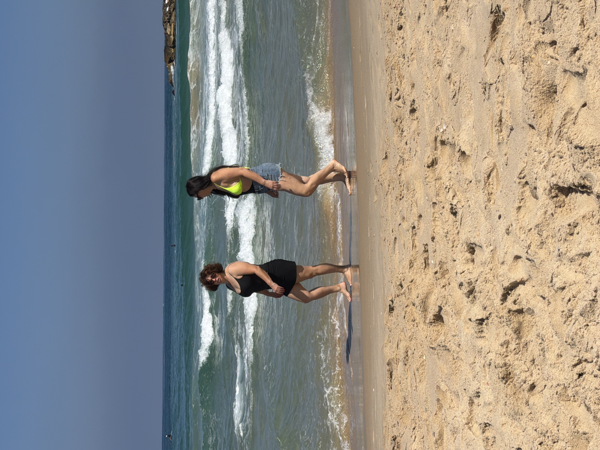 Two women are walking on the beach in Ashkelon, Israel. The woman on the left is wearing a black dress and has curly brown hair. She is looking towards the viewer. The woman on the right is wearing a neon yellow bikini top and denim shorts. She is looking to her left. The ocean is behind them with waves breaking on the shore. The sand in the foreground is light brown and textured. The sky is clear and blue.