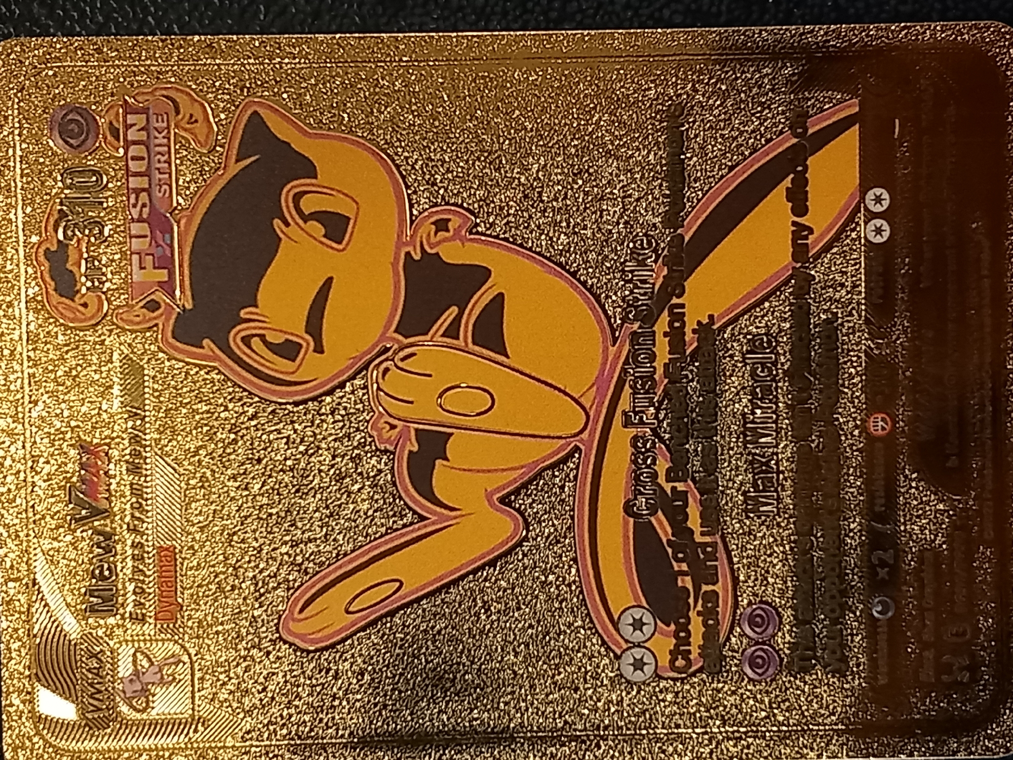 The image is a close-up, high-angle shot of a gold-foiled Pokemon card featuring Mew VMAX. The card is a "Fusion Strike" set card with a HP of 310. The artwork depicts Mew VMAX in a playful, stylized pose with its legs and arms extended. The card's text is mostly legible, describing its abilities, "Gross Fusion Strike" and "Max Miracle." The background of the card is a textured gold pattern, common for special edition Pokemon cards. The overall lighting is bright, highlighting the metallic sheen of the card. There are no other objects or people visible in the frame, and the setting is not discernible beyond the card itself.