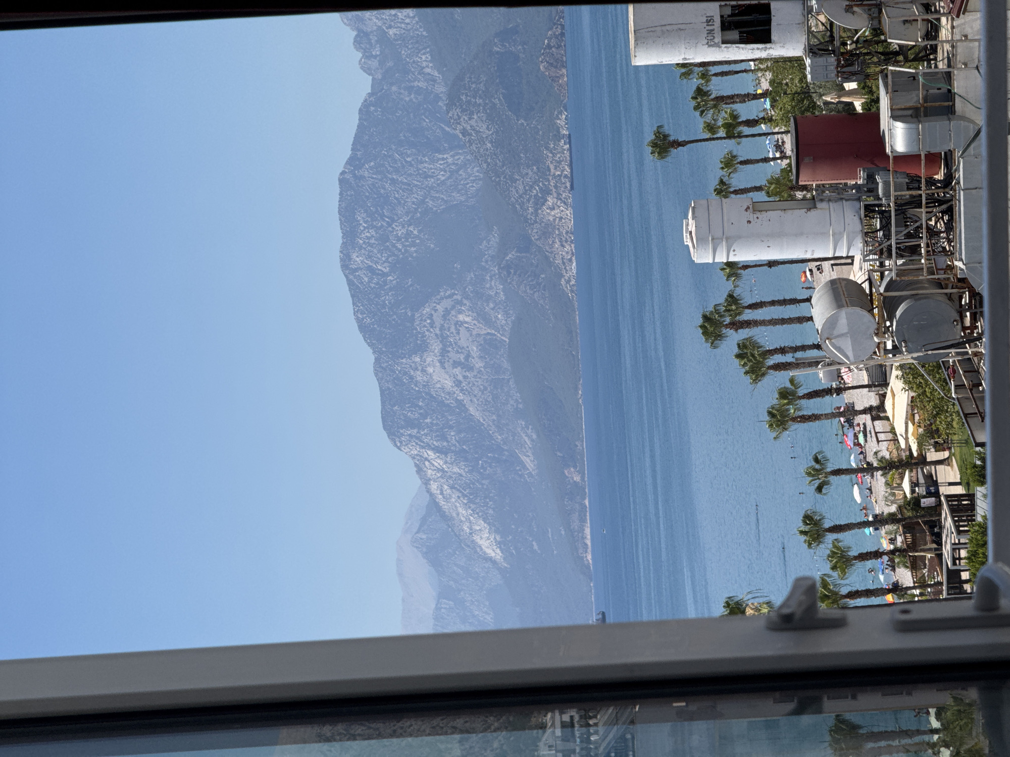 This media file depicts a view from a window overlooking a coastal scene. The primary subjects include a calm, blue sea, a steep, rocky mountain range in the background, and a sandy beach lined with palm trees. Scattered on the beach are numerous people enjoying the day, with many lying on towels and others swimming in the water. Beach umbrellas provide shade, suggesting a sunny day. Several structures, possibly part of a resort or beach facilities, are visible, including cylindrical tanks and metal scaffolding. The scene is captured from a slightly elevated perspective, looking out through a window frame. The clear blue sky indicates good weather, likely during daylight hours, possibly late morning or early afternoon. Reflection in the window shows the interior of the room from which the photo was taken. The overall mood is relaxed and typical of a seaside vacation. There is faint text visible on one of the white cylindrical structures, appearing to be Turkish: "GÜN İŞİ". The location context is identified as Konyaaltı, Turkey.