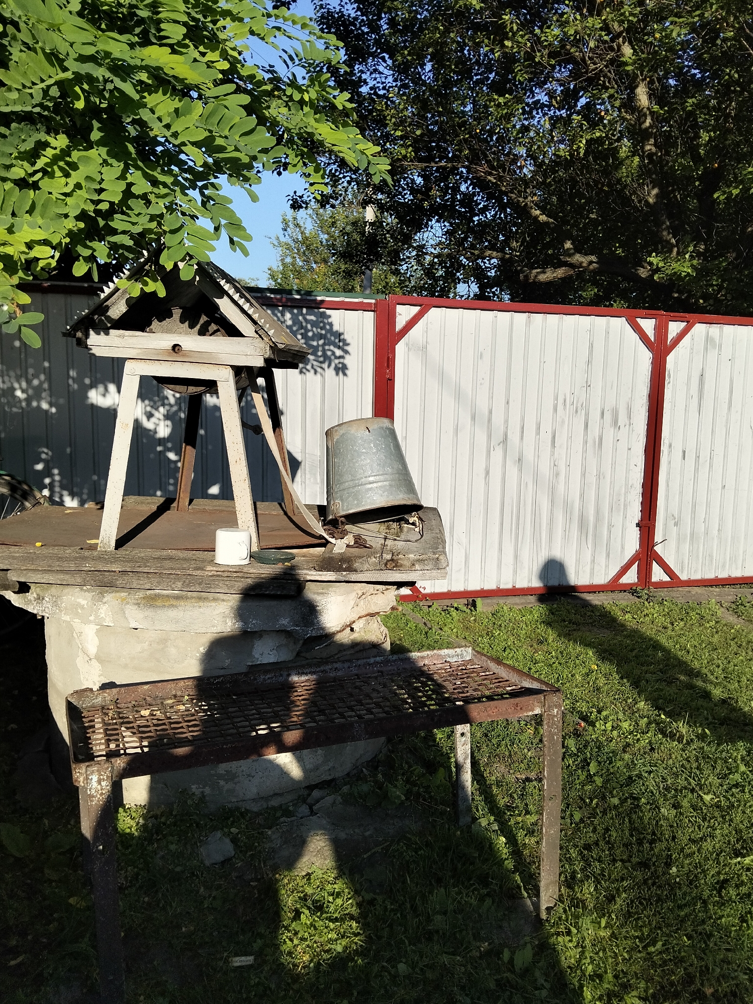 In Kliushnykivka, Ukraine, a well stands under a tree, its wooden roof casting a shadow. A metal bucket rests on the well's edge, next to a white mug. In the foreground, a rusty metal table is set up, its shadow falling on the grass. A white fence with red accents borders the property, with trees and a clear blue sky in the background.