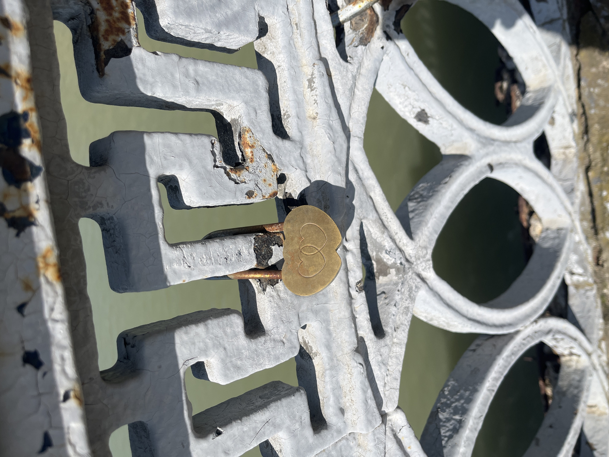 A close-up depicts a section of a gray, ornate metal fence with peeling paint and some rust, with a gold-colored love lock attached to the fence. The lock features two interlocking heart-shaped designs. The background shows a blurred expanse of light green water, suggesting the fence is situated near a body of water. The style of the fence is reminiscent of historical architectural details. Based on the presence of the love lock, this is likely a location where couples attach locks as a symbol of their affection. The location is Krasnodar, Russia, as love locks are a common practice in urban areas.