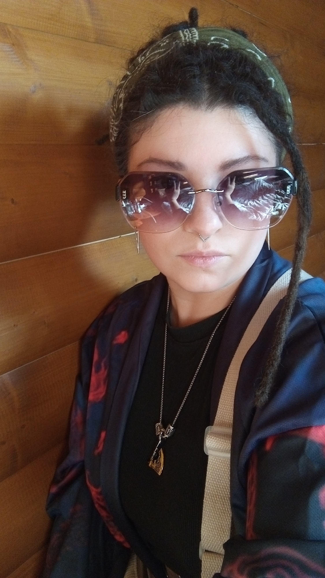 A woman with dark hair styled in dreadlocks and a green patterned bandana poses for a selfie against a wood-paneled wall. She wears large, semi-transparent sunglasses, a nose ring, and dangling earrings. She is dressed in a black top and a blue and red patterned kimono-style jacket, with a silver necklace featuring an axe pendant visible. A light-colored strap crosses her torso. The reflection in her sunglasses suggests an interior setting, possibly a restaurant or cafe in Юдино, Russia.