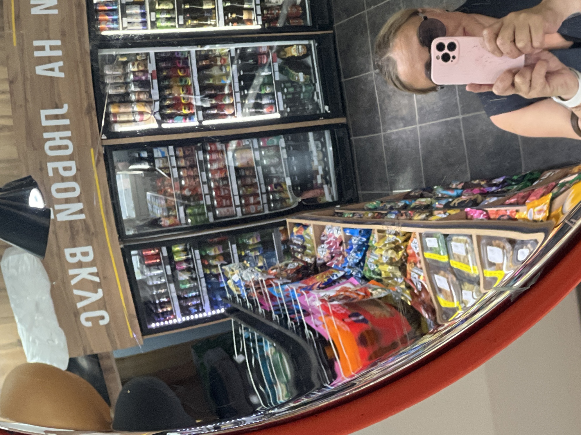 A person wearing sunglasses is taking a photograph of themselves in a convex mirror. The reflection includes shelves of snacks and refrigerated drinks, along with a sign reading "продукты ВКАС". The location is Chaikovsky, Russia.