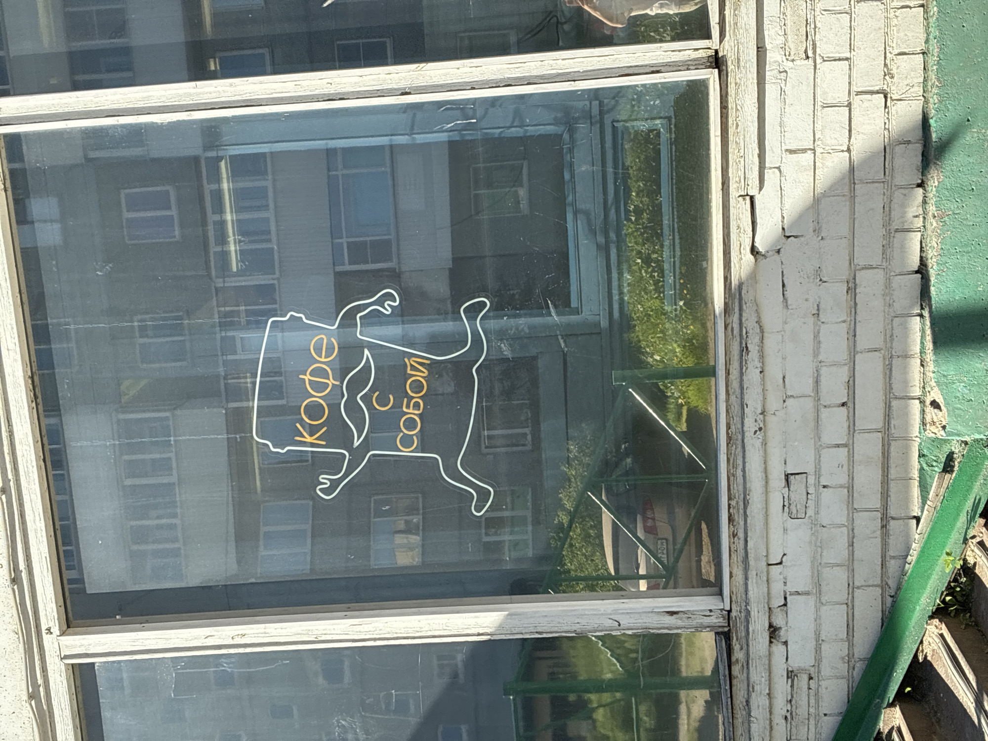 The image shows a window with a neon sign on it that reads "Кофе с собой," which translates to "Coffee to go." The sign depicts a stylized coffee cup with arms and legs running, and a mustache. The window reflects buildings and greenery outside. Below the window is a brick wall painted white, showing signs of wear and tear. There is also a green metal railing and a portion of a car visible through the window. The scene appears to be an exterior shot of a building, likely a cafe or coffee shop, in an urban environment.