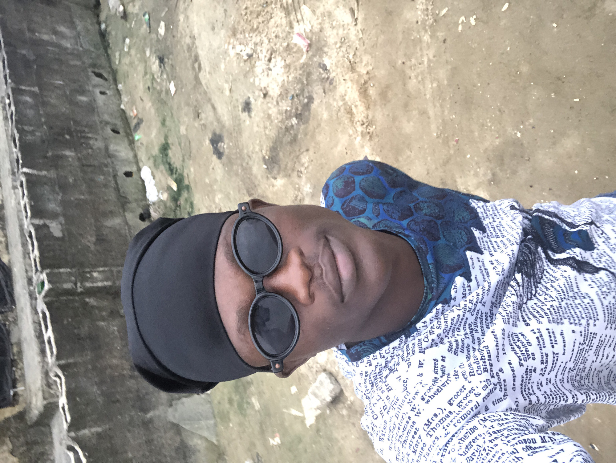 A young Black male, appearing to be in his late teens or early twenties, is captured in a high-angle, forward-facing selfie. He wears a black durag and large, oval-shaped dark sunglasses that obscure his eyes. His facial features are clearly visible, including his nose, lips, and jawline. He is dressed in a white t-shirt with a repeating pattern of black text, and a blue and black patterned shirt is visible beneath it. The background suggests an outdoor urban environment, likely in Ado, Nigeria. To the left and top of the frame, a weathered concrete wall, possibly part of a drainage system or a low-lying building, is visible. The ground is composed of dirt and rubble, with scattered debris such as plastic bags and paper, indicative of a less maintained area. The overall impression is a candid street portrait.