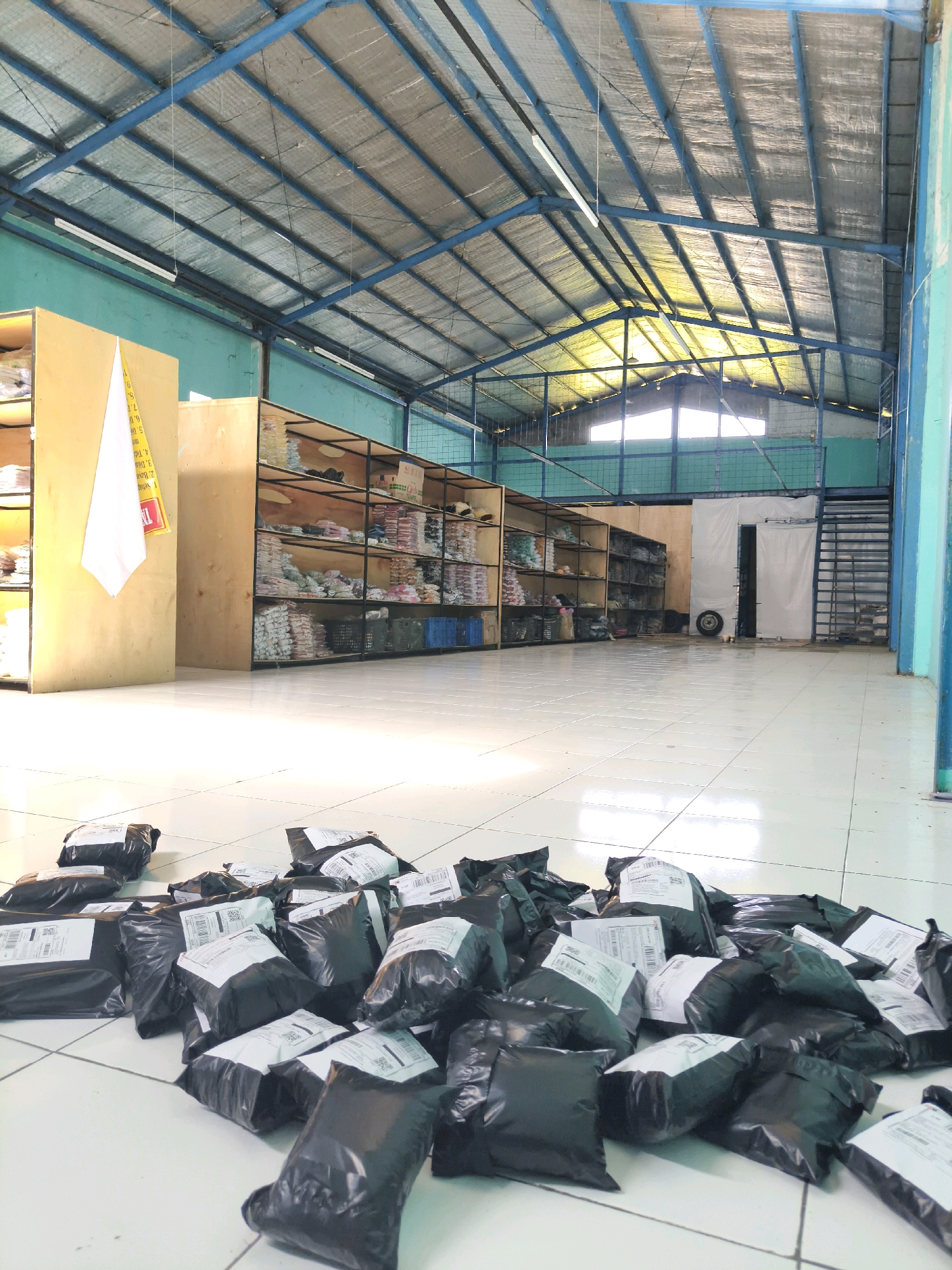 The image depicts the interior of a warehouse in Majasetra, Indonesia. The warehouse features a high, blue-trussed metal roof with silver insulation. Fluorescent lights are suspended from the ceiling. The walls are painted turquoise. Along the left side of the warehouse are tall, light-colored wooden shelves filled with neatly stacked items, predominantly clothing in plastic packaging. On the right side, similar shelving units are visible, also containing packaged goods. The floor is tiled in a white, grid pattern. In the foreground, a large pile of black plastic-wrapped packages, presumably ready for shipment, is scattered across the floor. These packages vary in size and some have white shipping labels attached. In the background, a large white structure, possibly a temporary partition or a roll-up door, is present. A spare tire rests on the floor next to it. Further to the right, a metal staircase leads to an upper level. The overall scene suggests an active logistics or distribution facility. There are no people visible in the image.
