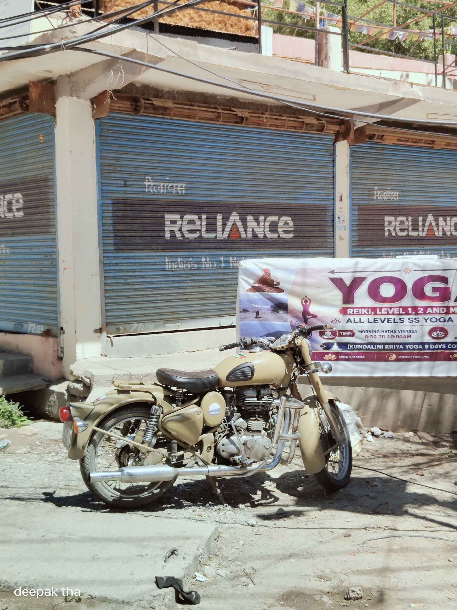 An outdoor, eye-level, full shot captures a tan-colored motorcycle parked on a dirt road in Leh, India. The motorcycle is the primary subject, facing slightly left, with its front wheel turned. Behind the motorcycle, a beige banner advertises "YOGA" with details on classes like "REIKI, LEVEL 1, 2 AND MORE" and "MORNING HATHA VINYASA 8:30 to 10:00 AM." The banner is displayed in front of a building with blue corrugated metal shutters. Text on the shutters reads "RELIANCE" in white lettering with an orange outline. The building's facade features exposed concrete beams and pillars, and the roof is a weathered, corrugated material. Overhead, electrical wires crisscross the frame, and prayer flags are visible on a balcony in the background. The ground is a mixture of dirt, gravel, and cracked concrete, with shadows cast by the motorcycle and overhead structures. The scene is lit by bright sunlight, creating strong contrasts and shadows. A watermark with the text "deepak tha" is present in the lower left corner of the image.