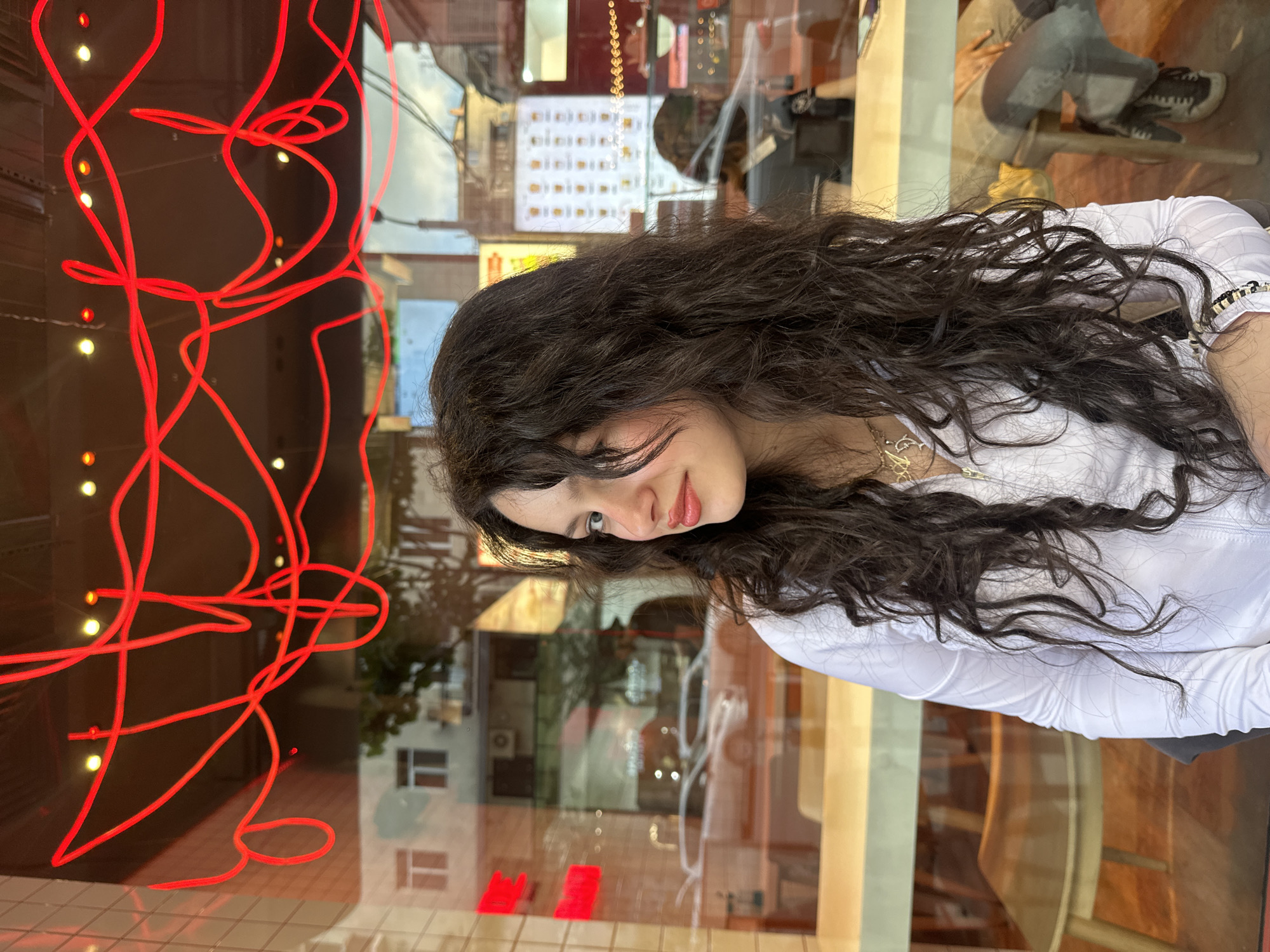 A young woman with long, dark curly hair, wearing a white long-sleeved shirt and a gold necklace, is sitting indoors near a window. She is looking downwards with a slight smile. Behind her is a window with reflections of the street. There is a red neon light design on the window. Outside, a building, possibly an apartment complex, is reflected, and so is a view of the interior of the establishment: menu or product displays, other patrons, and furniture. Based on the reflection of the exterior elements, specifically the apartment building style and window designs, along with the style of the interior and the availability of visible menu/display items, it's plausible that this is Yerevan, Armenia.