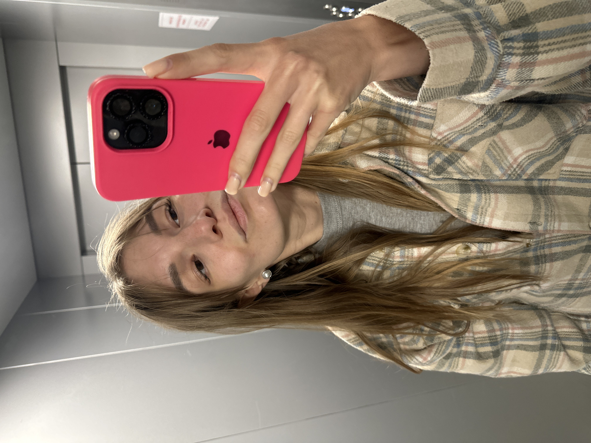 A young woman takes a mirror selfie in an indoor setting, likely an elevator or a room with reflective walls. The image is oriented sideways due to the selfie angle.
The primary subject is a woman with long, light brown hair and a neutral expression, visible from the chest up. She is wearing a light grey top layered with a loose-fitting plaid shirt or jacket in shades of beige, blue, and light pink. A simple, pearl-like stud earring is visible in her right ear.
She holds a hot pink-cased Apple iPhone in her right hand, positioned prominently in the foreground. The phone's rear camera module, featuring three large lenses and a LiDAR sensor, is clearly visible, along with the distinct Apple logo on the case. Her hand shows well-maintained, light-colored manicured nails.
The background consists of smooth, reflective, light gray panels, characteristic of an elevator interior or a modern hallway with mirrored surfaces. In the upper right corner of the reflected background, a blurry rectangular sign with indistinct white text on a red background can be vaguely seen, but no specific words are legible. The lighting is uniform and artificial, suggesting an indoor environment.
The activity is a self-portrait taken using a mirror reflection. The overall impression is casual and personal, captured in Chaikovsky, Russia.