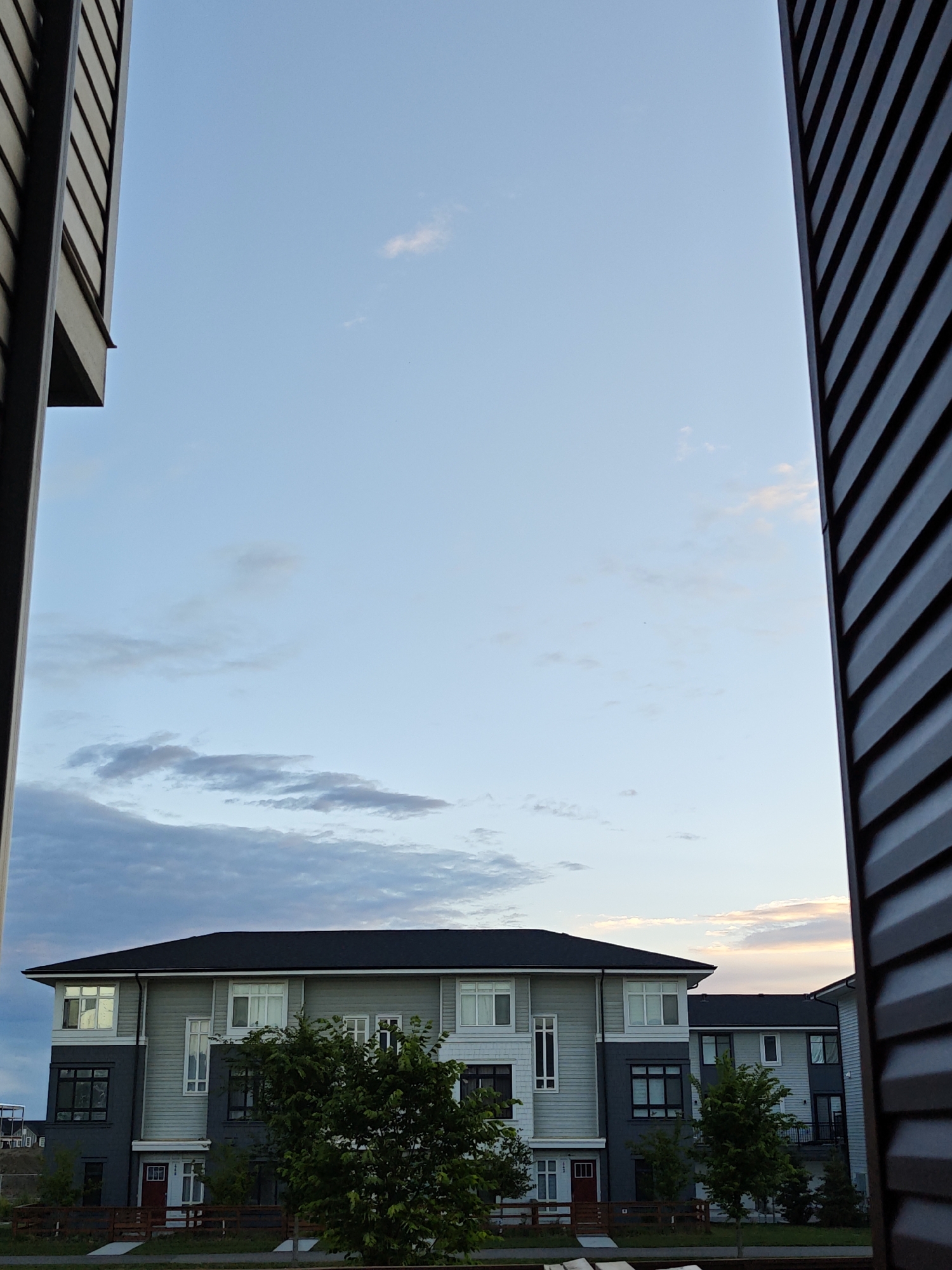 The image depicts a residential area with modern townhouses, likely in the city of Calgary, Canada. The sky is a pale blue with scattered clouds, tinged with soft hues of pink and orange from what appears to be the setting sun. The townhouses are light grey and dark grey, with black roofs and white-framed windows. Some of the windows reflect the sky. In front of the townhouses, there are green trees and a wooden fence. The scene is framed by the dark, textured siding of two buildings on either side of the image, suggesting the photo was taken from a window or balcony. The lighting suggests it is either early morning or late evening. There are no people visible in the image.