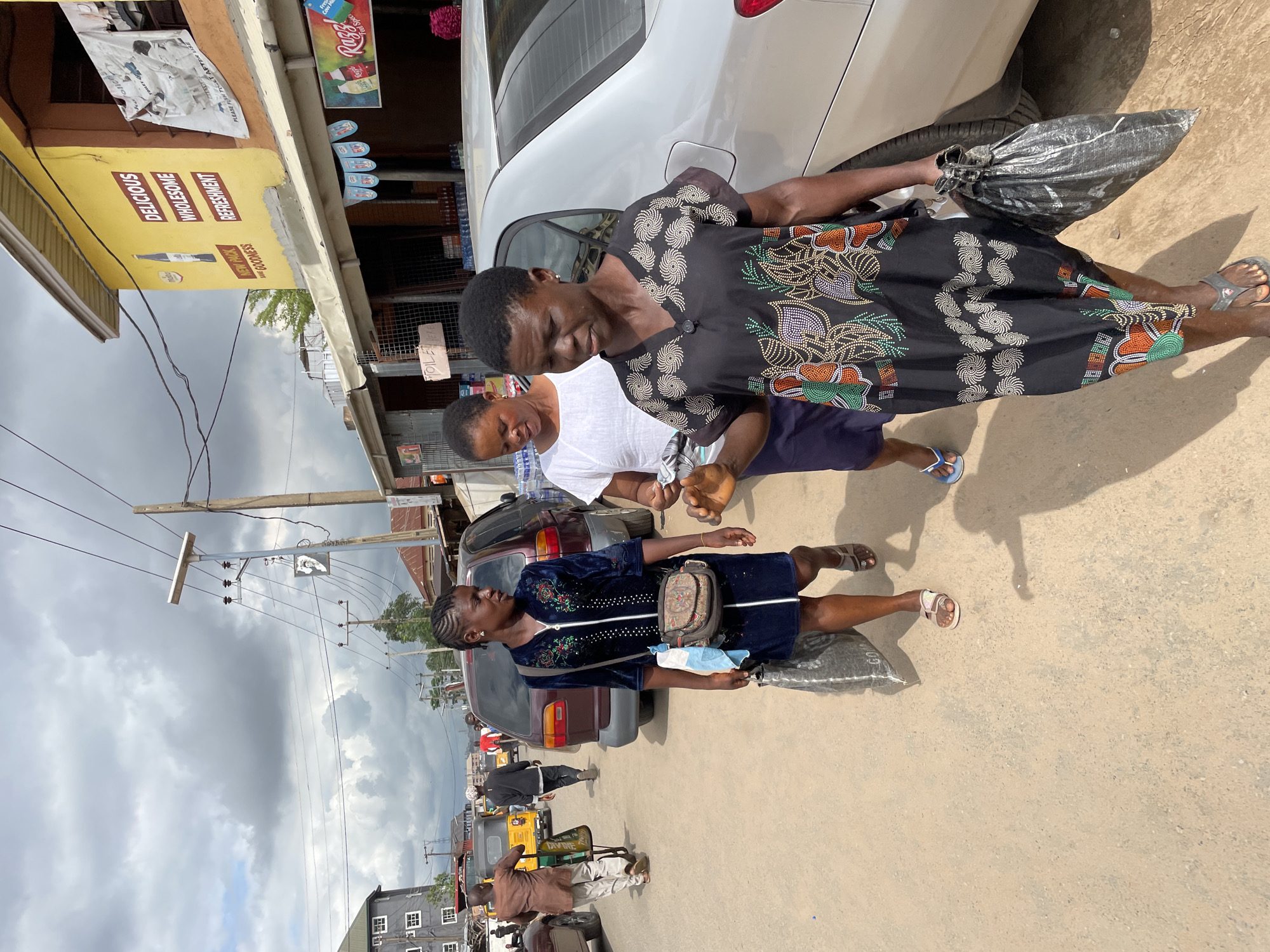 The image shows a street scene in Onitsha, Nigeria. Three women are visible in the foreground, two next to a silver car and the other further down the street. One woman is wearing a black dress with a floral pattern and is holding a gray bag. Another is wearing a white shirt and a dark skirt. The third woman is wearing a dark blue dress with a small purse. There are other vehicles in the street, including cars and a tricycle taxi (keke). There is a building on the left side of the frame with a yellow sign that says "DELICIOUS, WHOLESOME REFRESHMENT." Above the sign, the text states, "NEW PACK GOODNESS". Electrical poles and wires run along the top of the scene. The sky is cloudy.