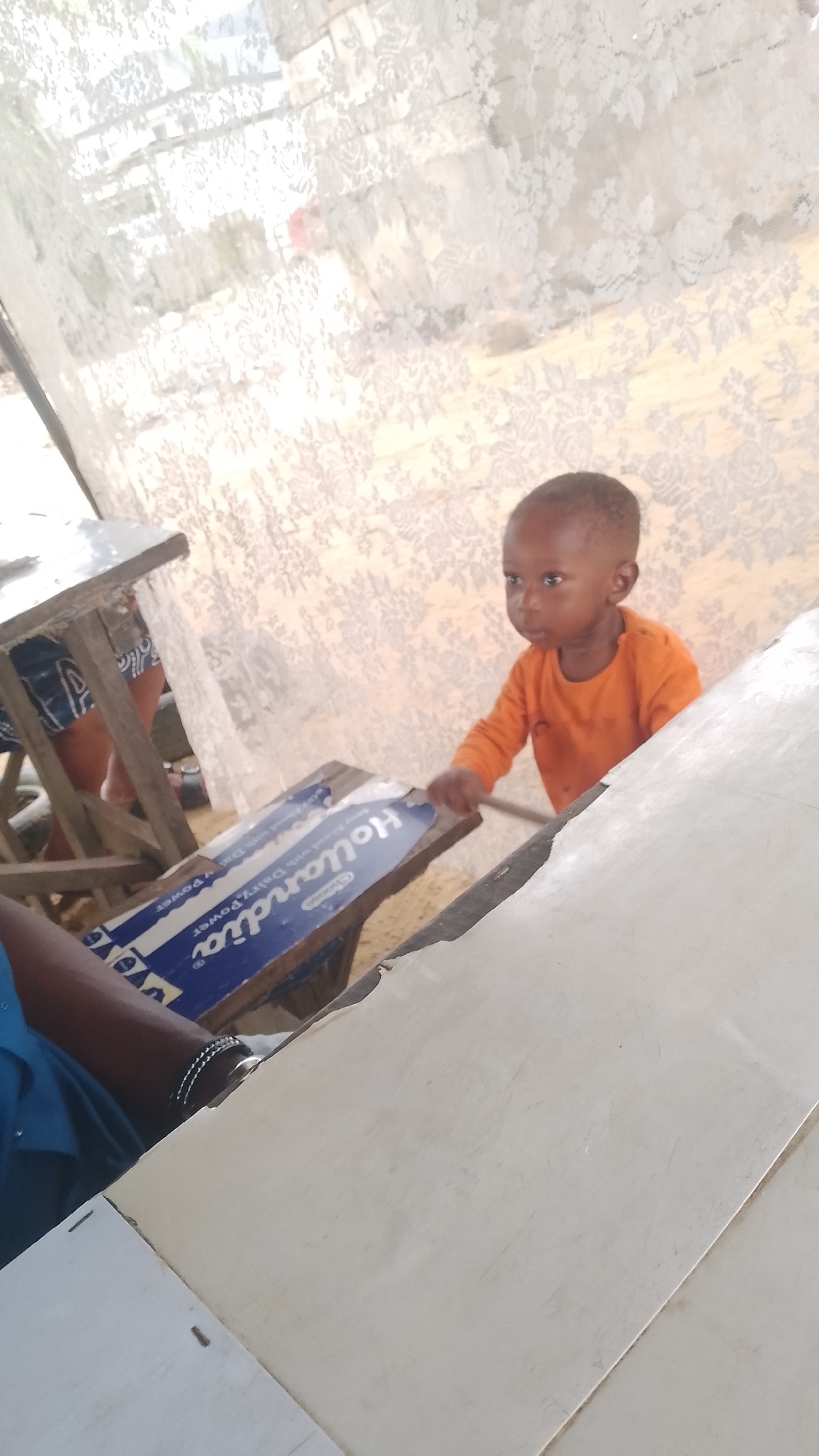 A child in an orange shirt stands next to a table in what appears to be a building in Effurun, Nigeria. The table is adjacent to one with a "Hollandia Dairy Power" advertisement attached. A person wearing a blue shirt is seated nearby with their arm and watch visible. Light filters through a patterned curtain or window covering in the background.