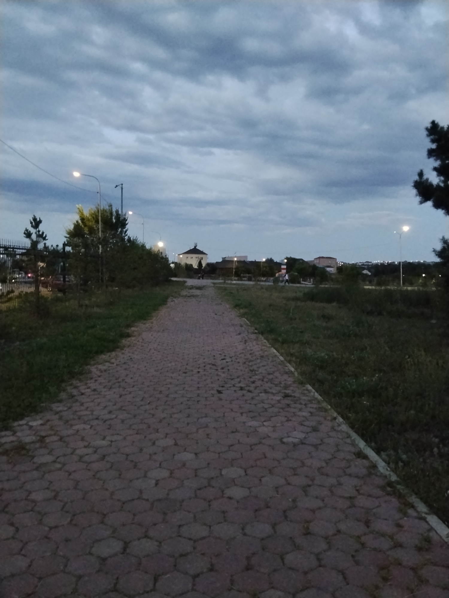 A paved pathway, constructed from hexagonal-shaped blocks, leads into the distance. The path is bordered by grass on the right and a mixture of grass, shrubs, and trees on the left. In the background, several buildings, including houses and what appears to be a lighthouse-like structure, are visible. Streetlights are illuminated along the pathway and in the distance, suggesting it is dusk or early evening. A fence is present on the far left of the image. The sky is overcast with gray clouds. Two individuals are walking on the pathway in the mid-ground. The image was taken in the city of Kokshetau, Kazakhstan.