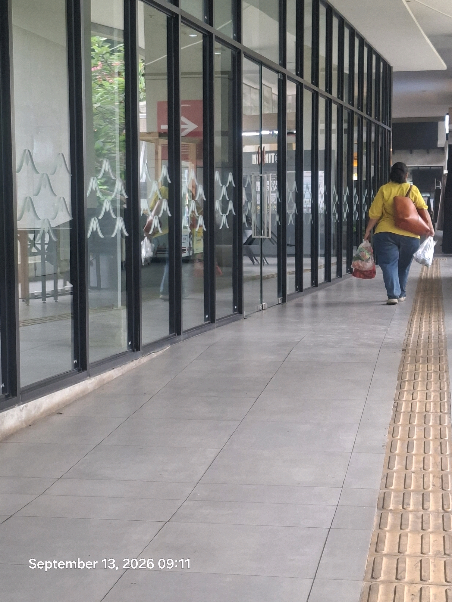 A person with dark hair, wearing a bright yellow shirt and blue jeans, walks away from the camera carrying two plastic bags and an orange purse over their right shoulder. The person is walking on a tiled sidewalk with a textured yellow tactile paving strip to their right. To the left of the sidewalk is a glass-paneled building with black frames. Inside the building, reflections show greenery and an arrow sign. The ground outside the building is also tiled. A timestamp at the bottom left of the image reads "September 13, 2026 09:11". The scene appears to be in Central Jakarta, Indonesia, based on the context provided.